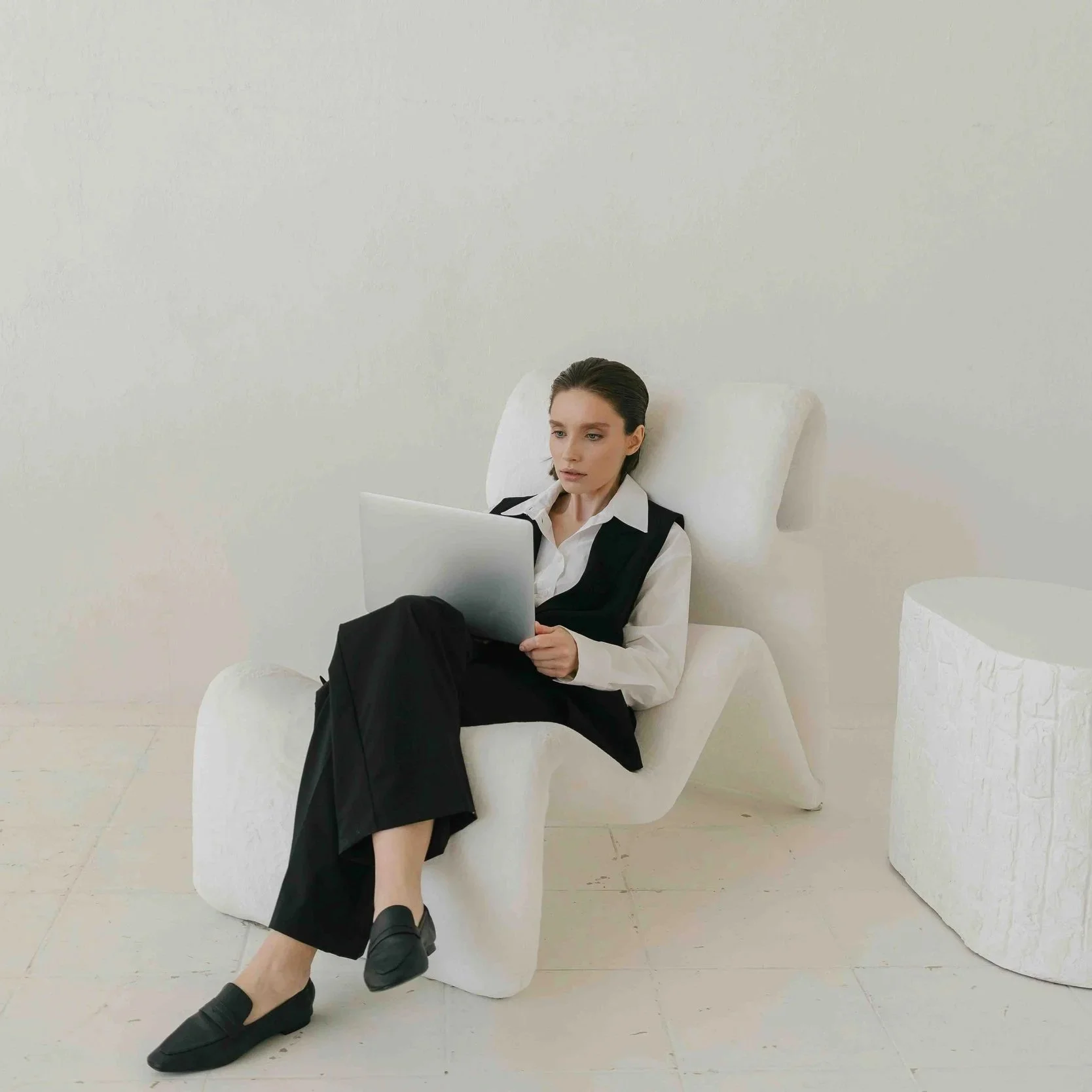 Woman in black and white outfit sitting in a modern white armchair, working on a laptop, with a potted plant nearby.