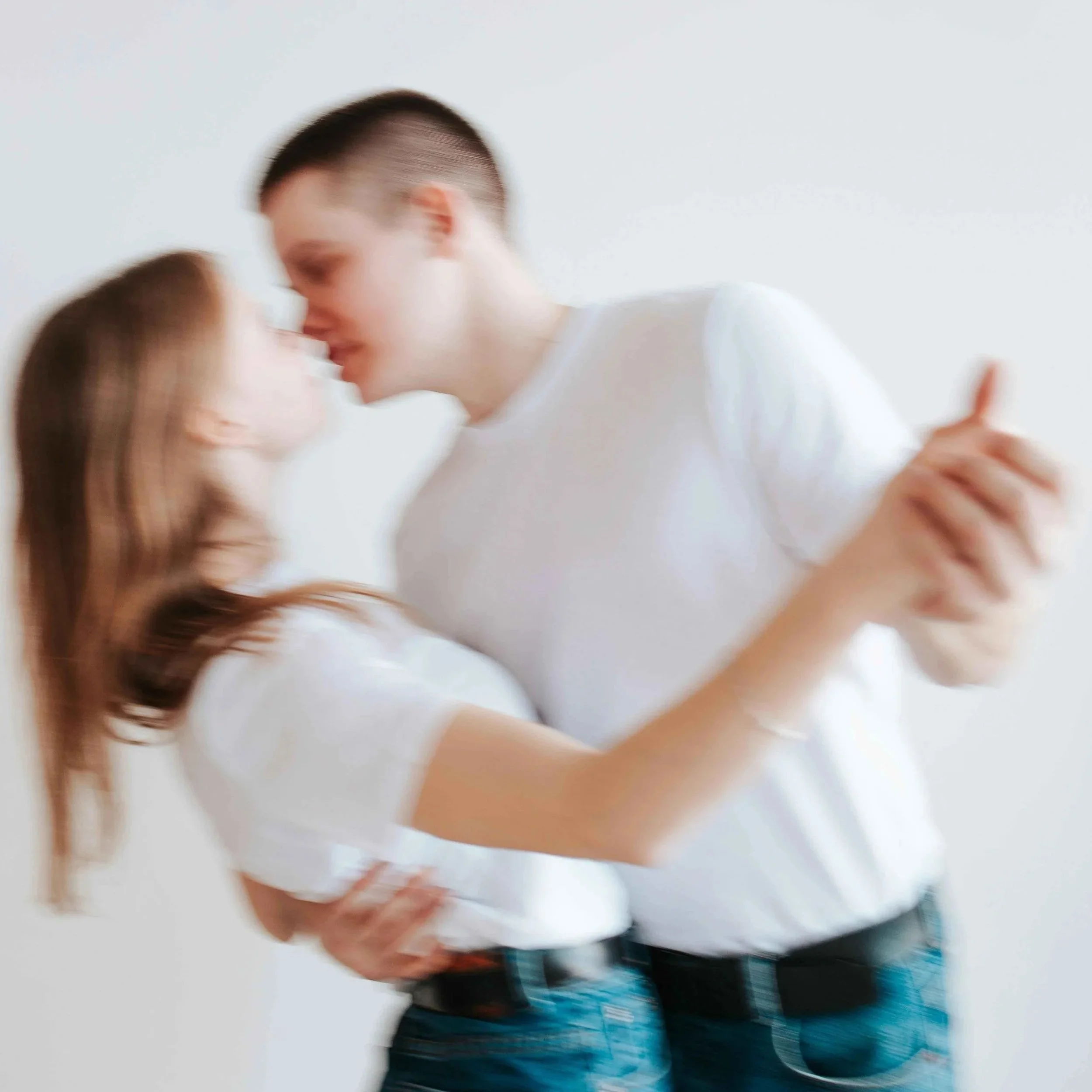 A blurry photo of a young couple dancing closely, about to kiss, in casual white t-shirts and dark jeans against a plain white background.