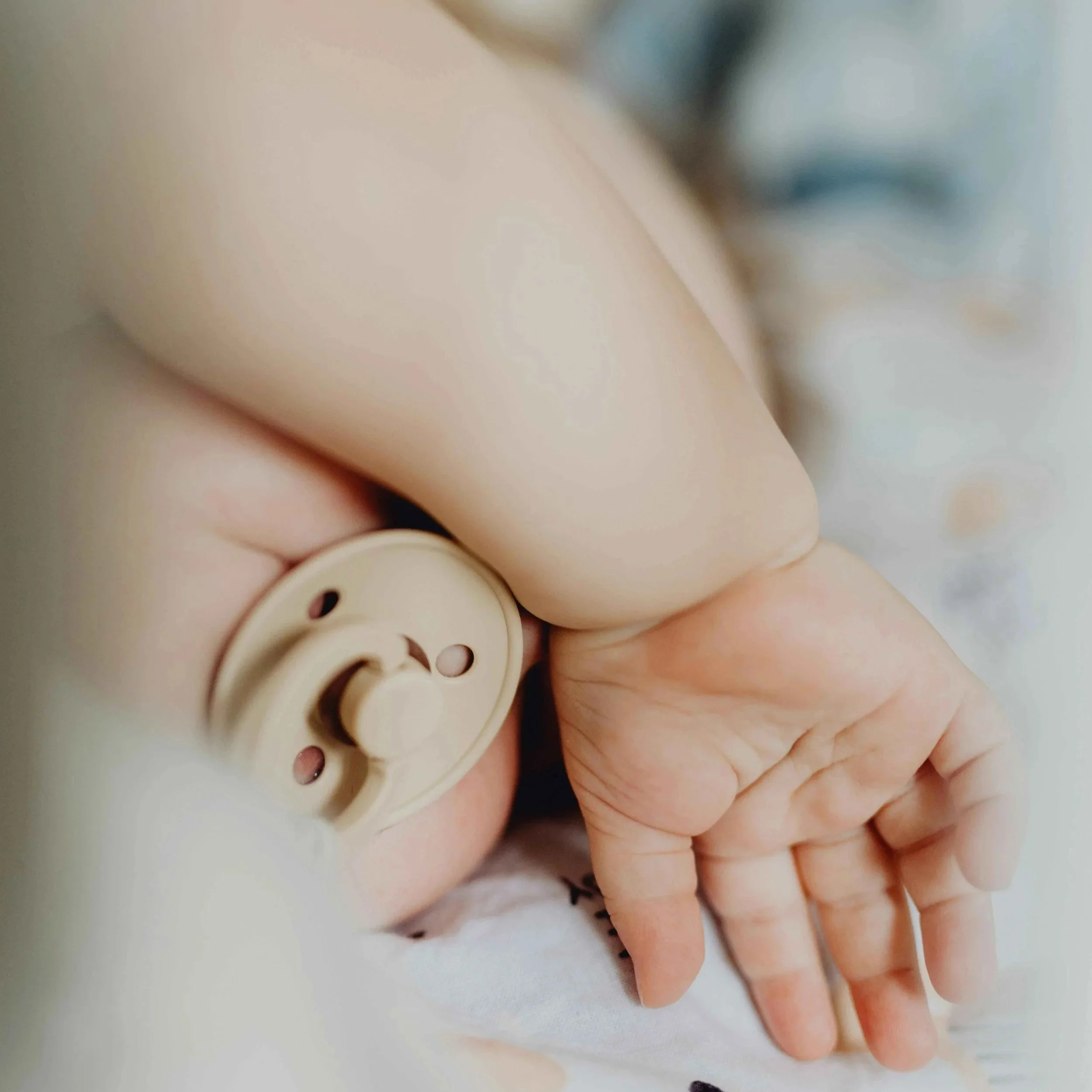 Close-up of a newborn baby's hand gripping a beige hospital identification bracelet, with the baby lying on a blanket and an adult's arm and part of the baby's face slightly blurred in the background.