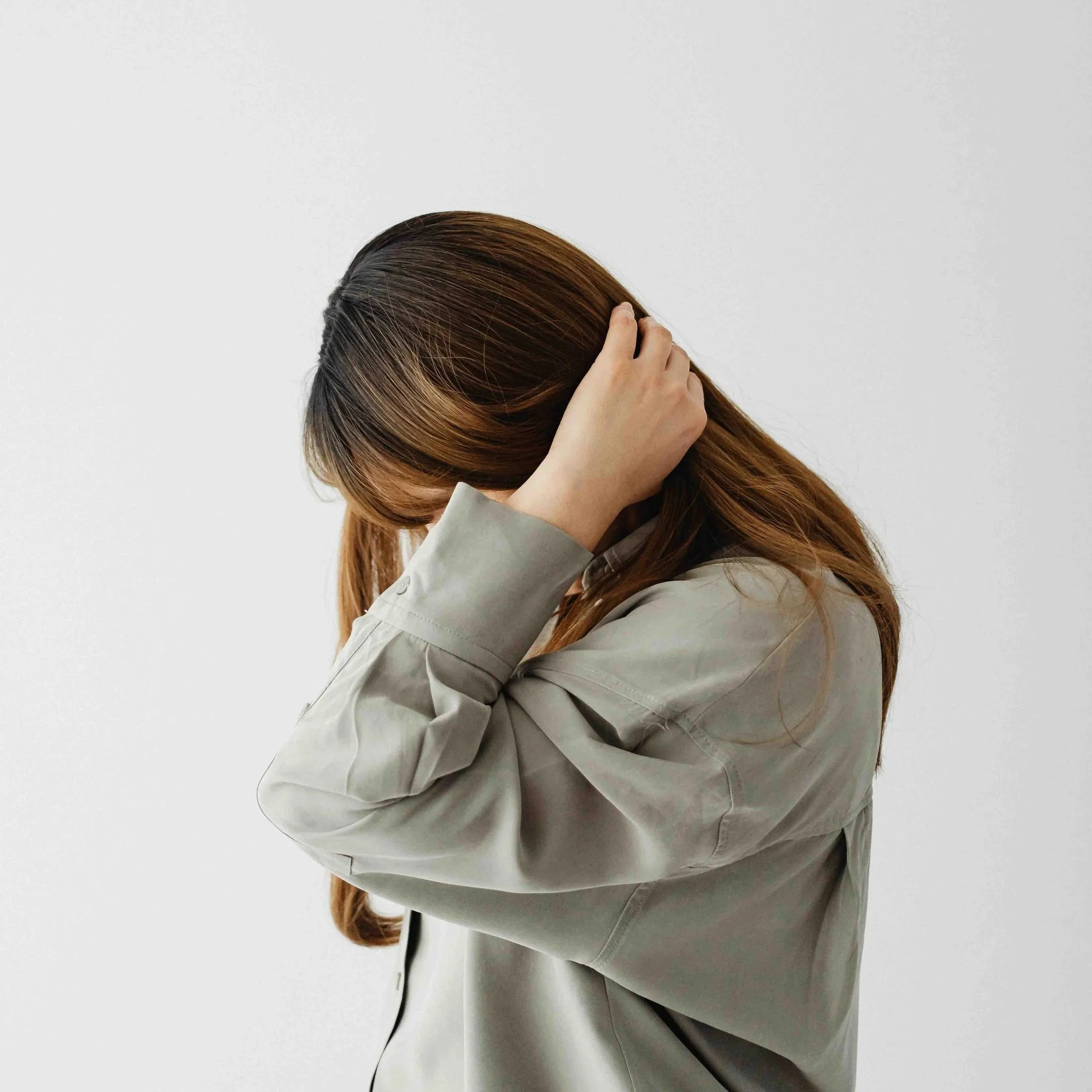 A woman with brown hair wearing a beige jacket, holding her head with both hands against a plain white background.