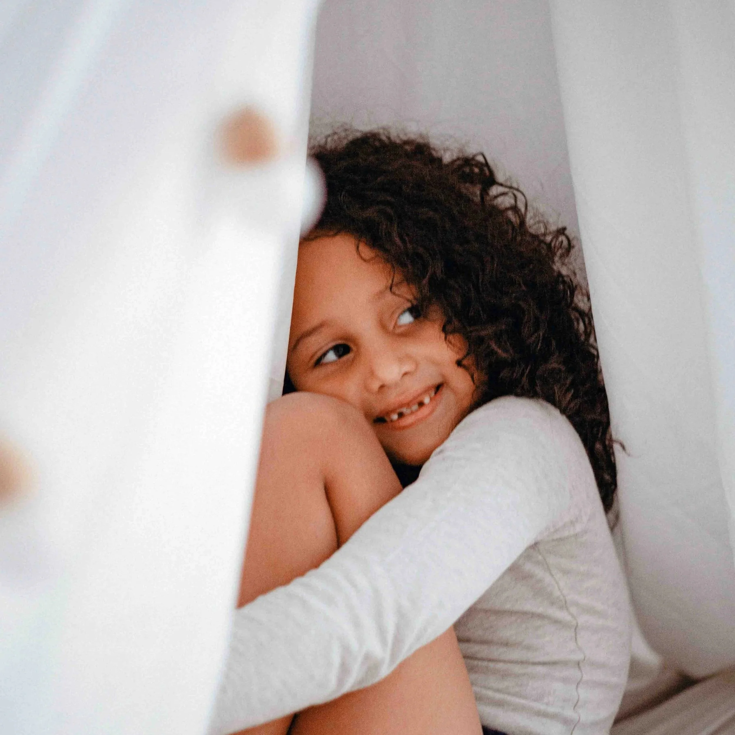 A young girl with curly hair smiling while sitting on a bed, hugging her knees, with a white pillow and curtains around her.