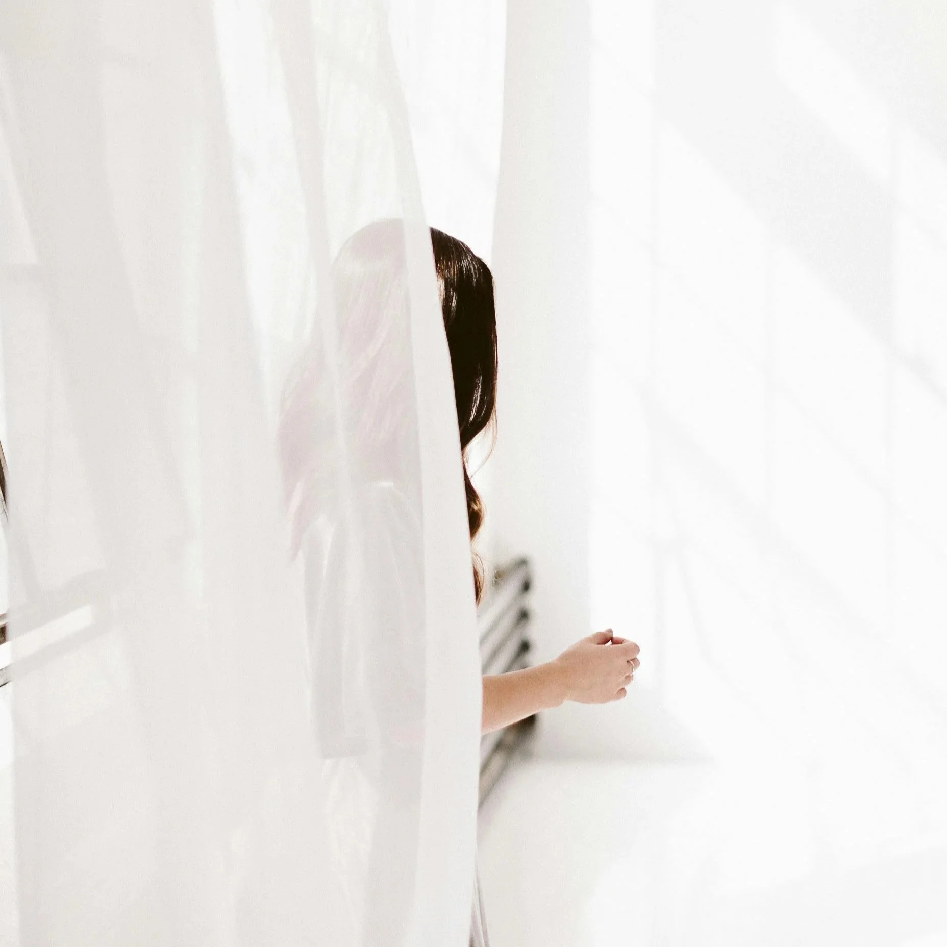 Person with long dark hair standing behind a semi-transparent white curtain in a bright, white room, with only the arm and hand visible.