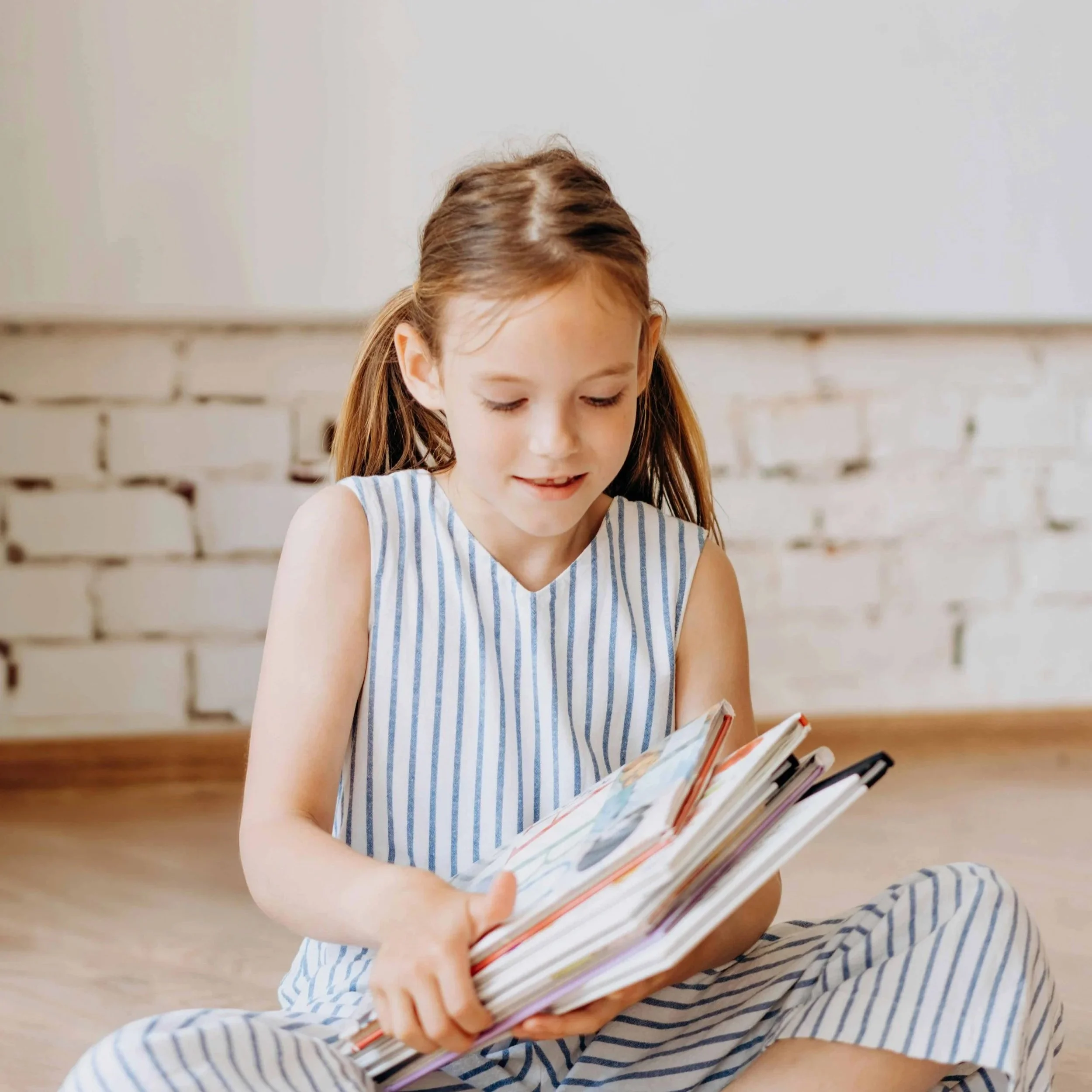 A young girl with long brown hair sitting cross-legged on a wooden floor holding books.