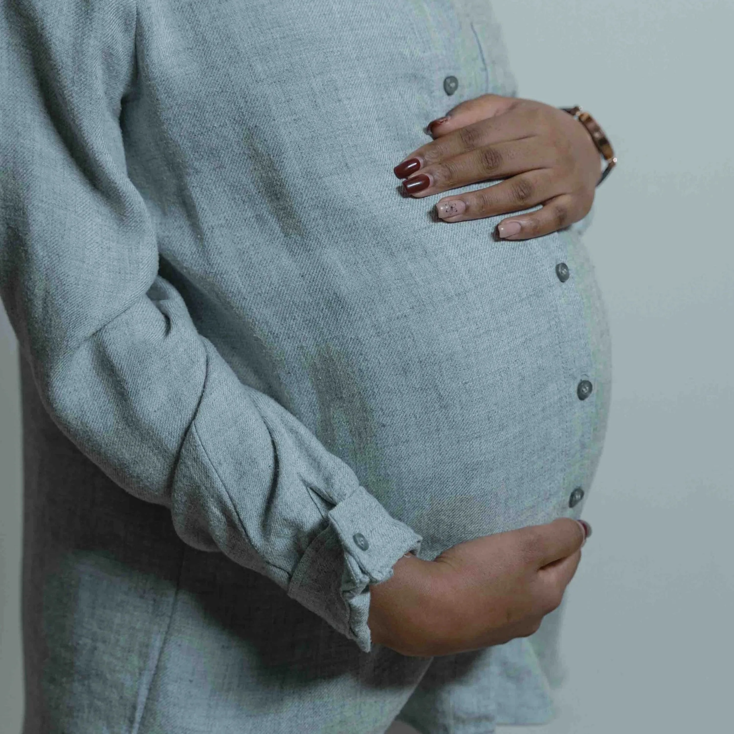 A pregnant woman wearing a gray button-up shirt, gently cradling her baby bump with her right hand on top and her left hand underneath.