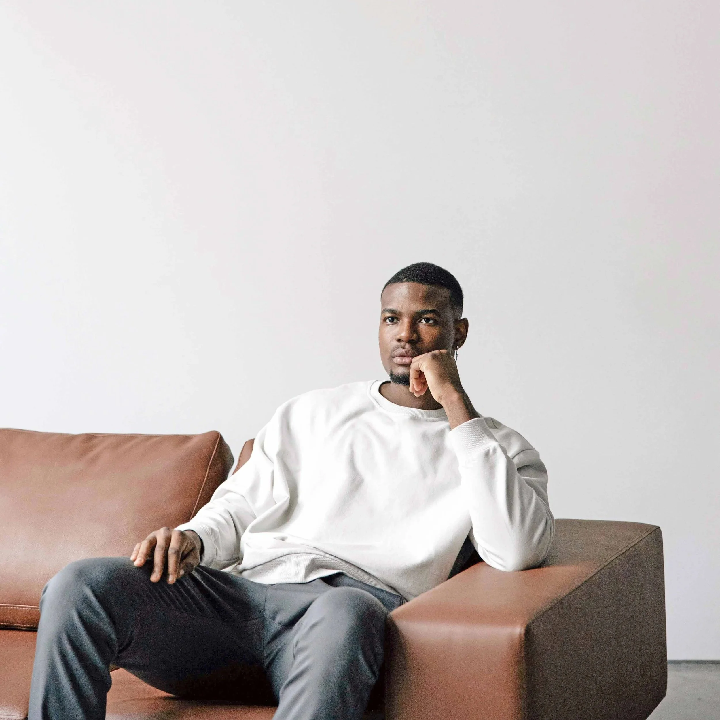 A young man with short dark hair, sitting on a tan leather sofa in front of a plain white wall, wearing a white sweatshirt and gray pants.