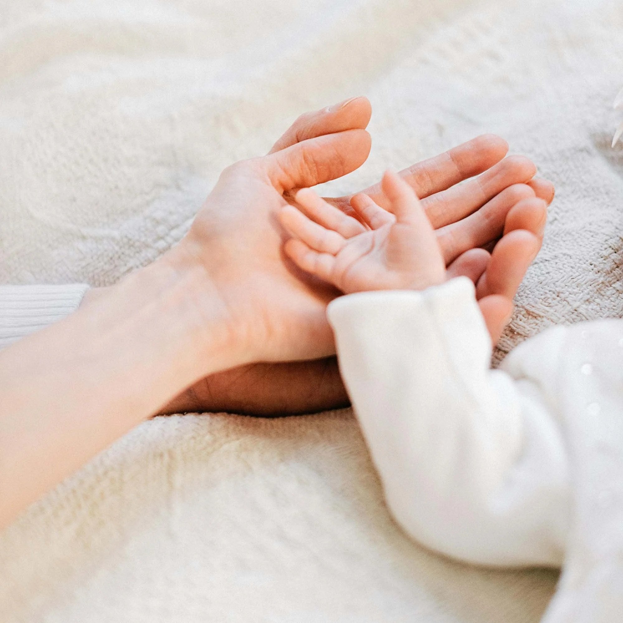 Close-up of an adult and baby holding hands on a cream-colored textured blanket.
