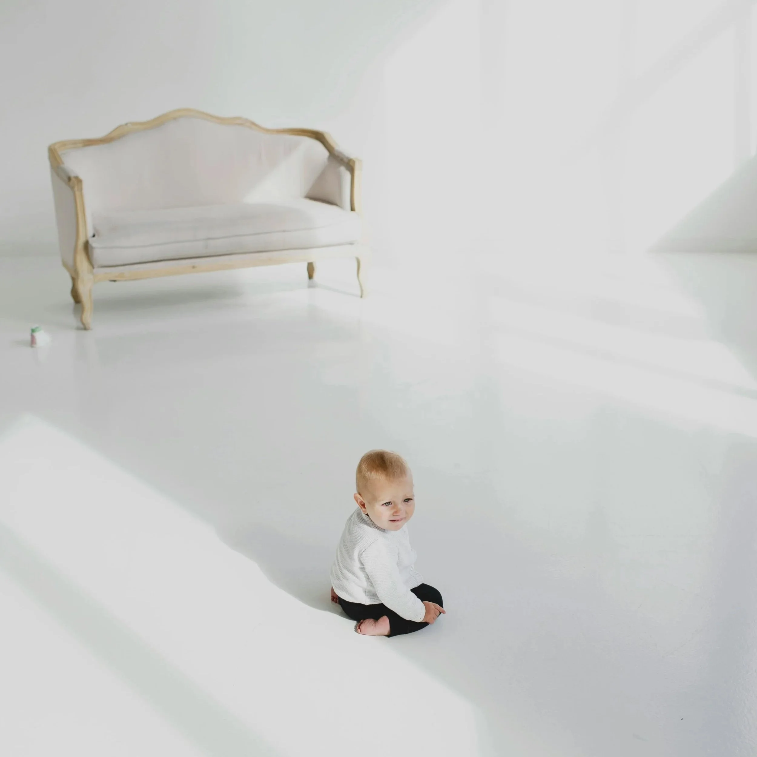 Child sitting on a white floor in a bright, minimal room with a white vintage sofa in the background.