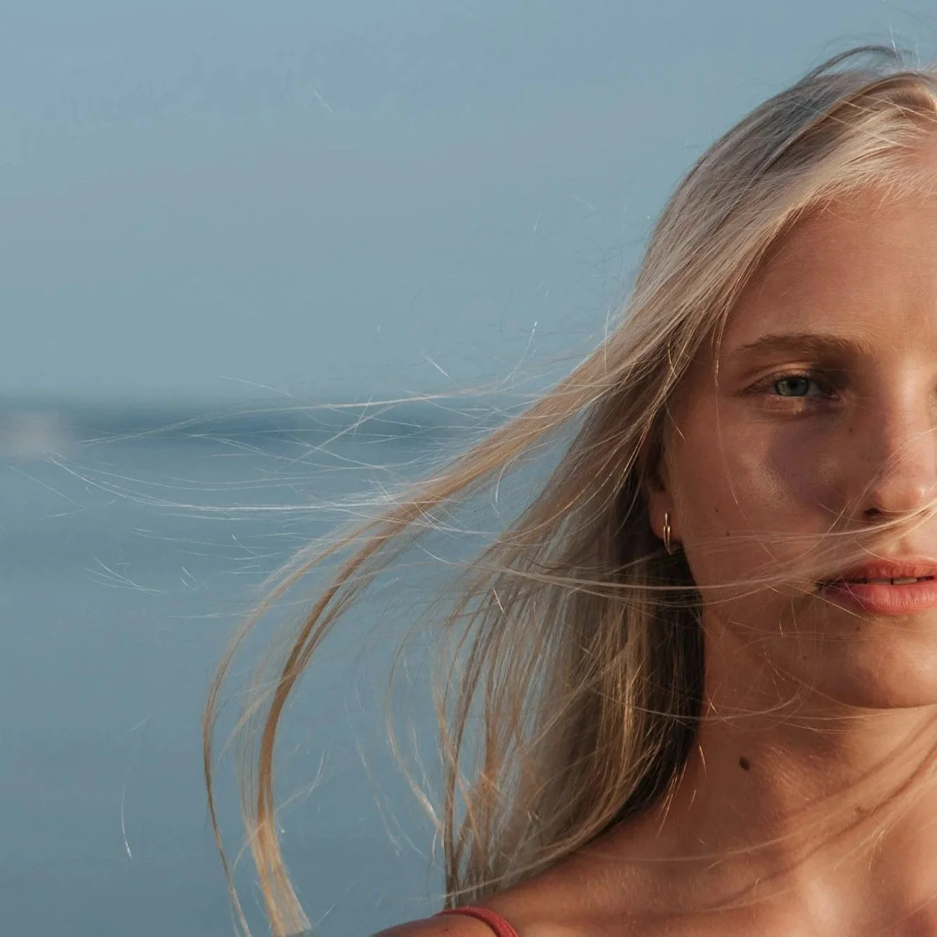 Close-up of a smiling blonde woman with windblown hair, wearing a sleeveless top, against a calm sea or lake background.