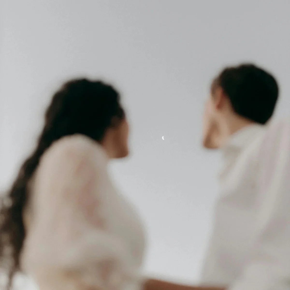 Two people sitting together looking at a crescent moon in a soft minimal sky.
