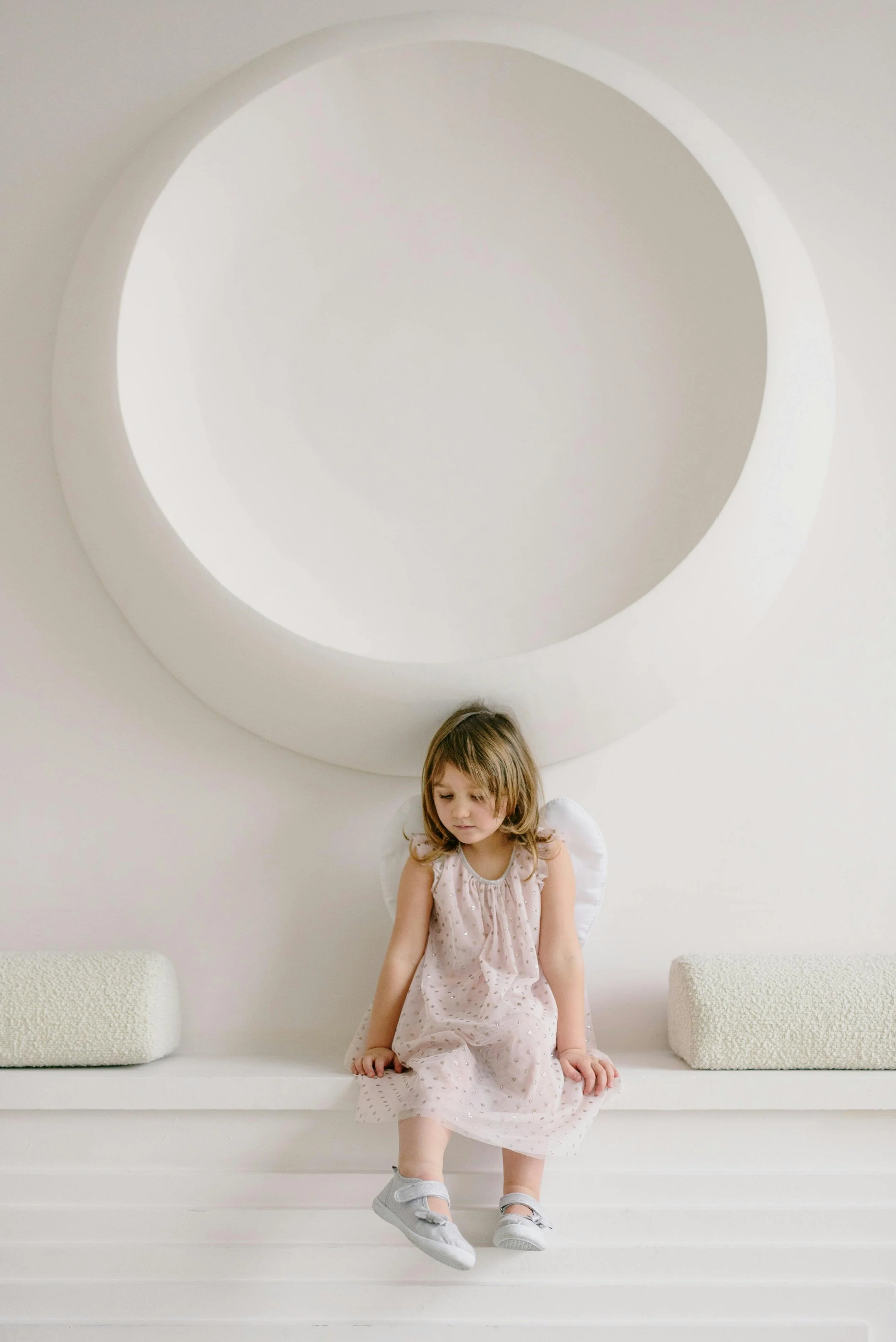 Little girl sitting beneath a large circular wall niche in a soft neutral interior.