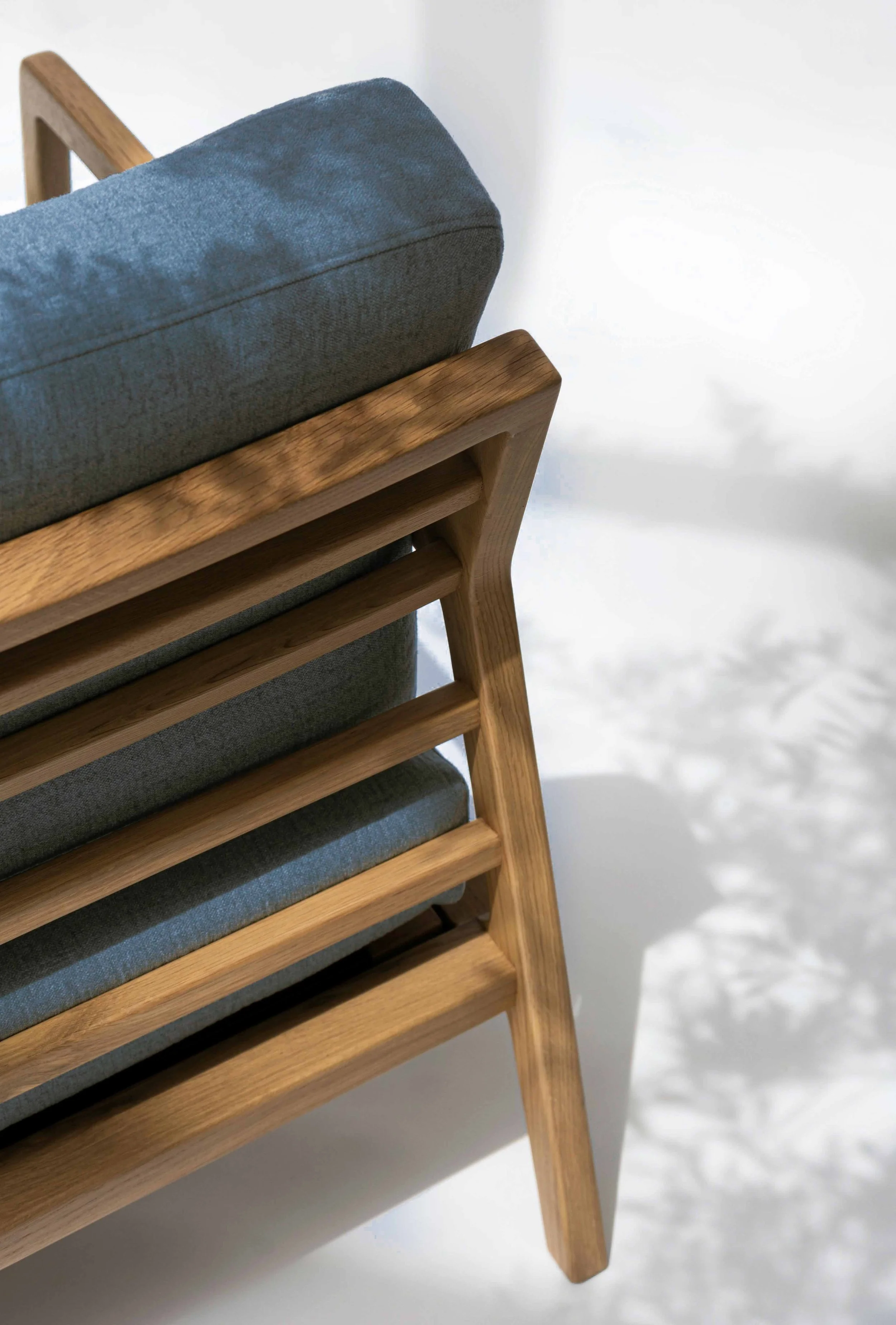 Minimalist wooden chair with blue cushion photographed in sunlight casting soft shadows on a white wall.