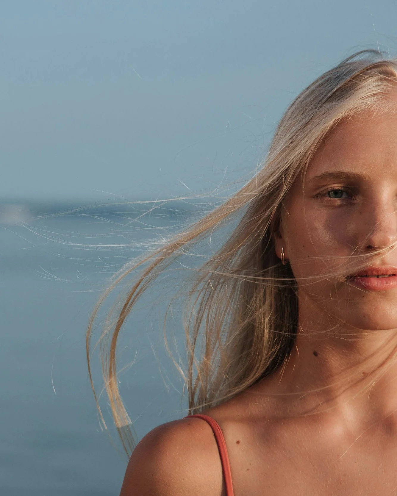 Close-up portrait of a blonde woman with wind-blown hair standing near water.