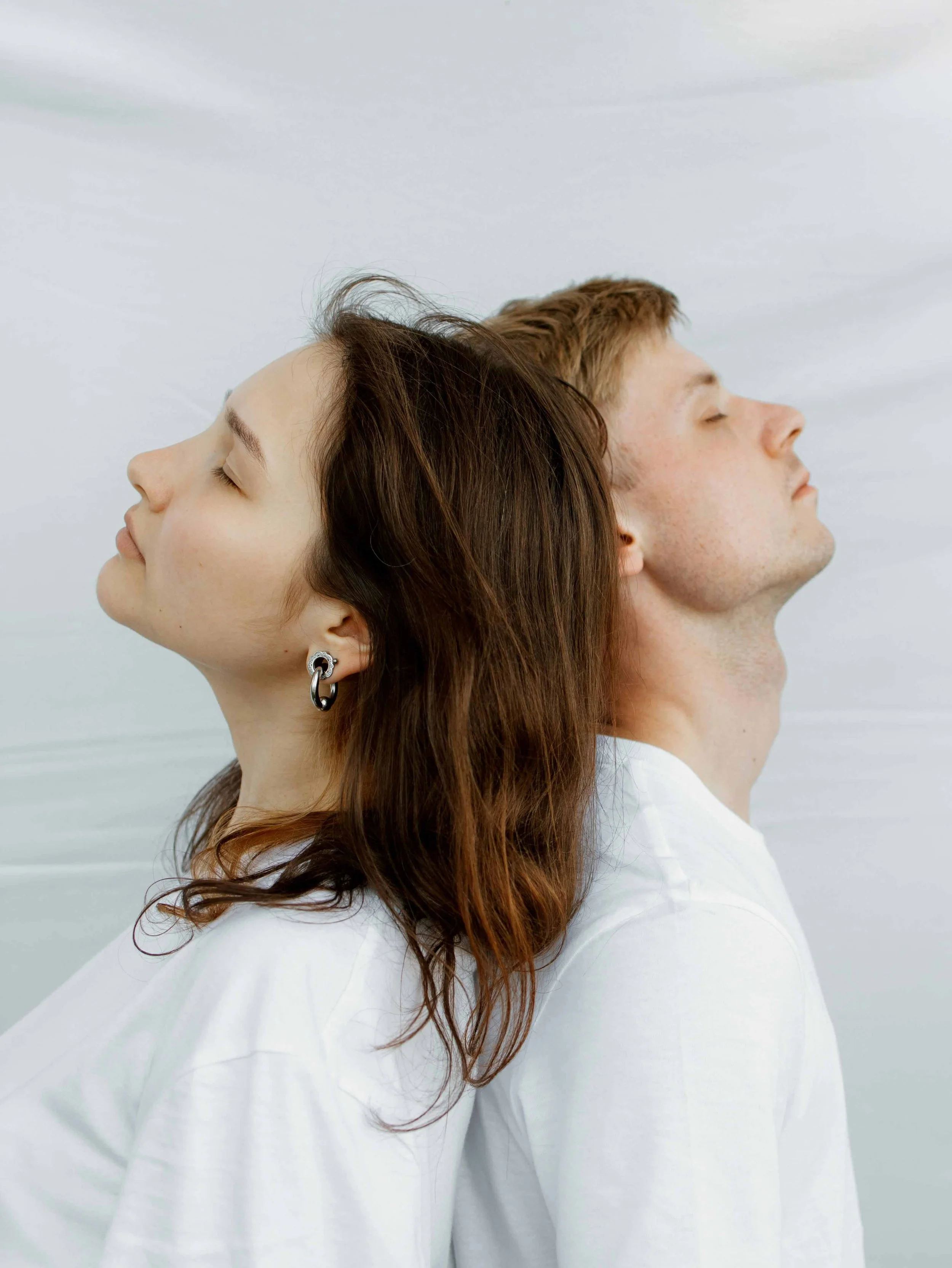 Two people standing back to back with eyes closed against a soft neutral background.