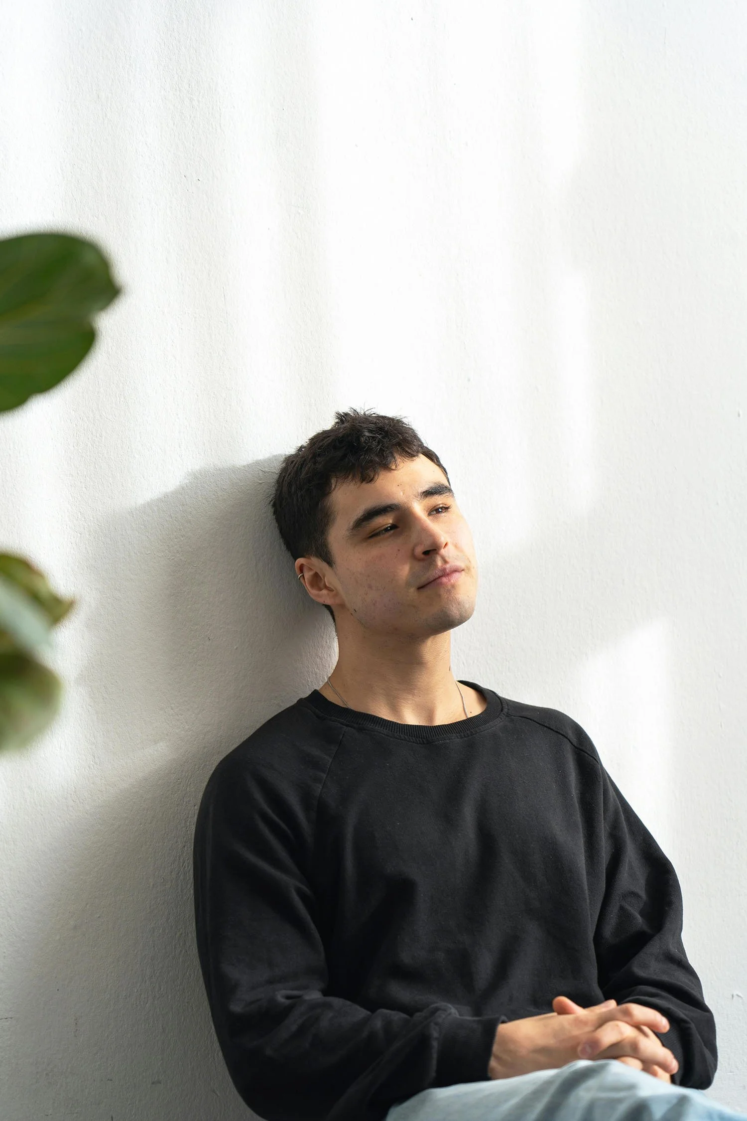 Young man in a black sweater sitting against a wall with hands clasped, looking reflective.