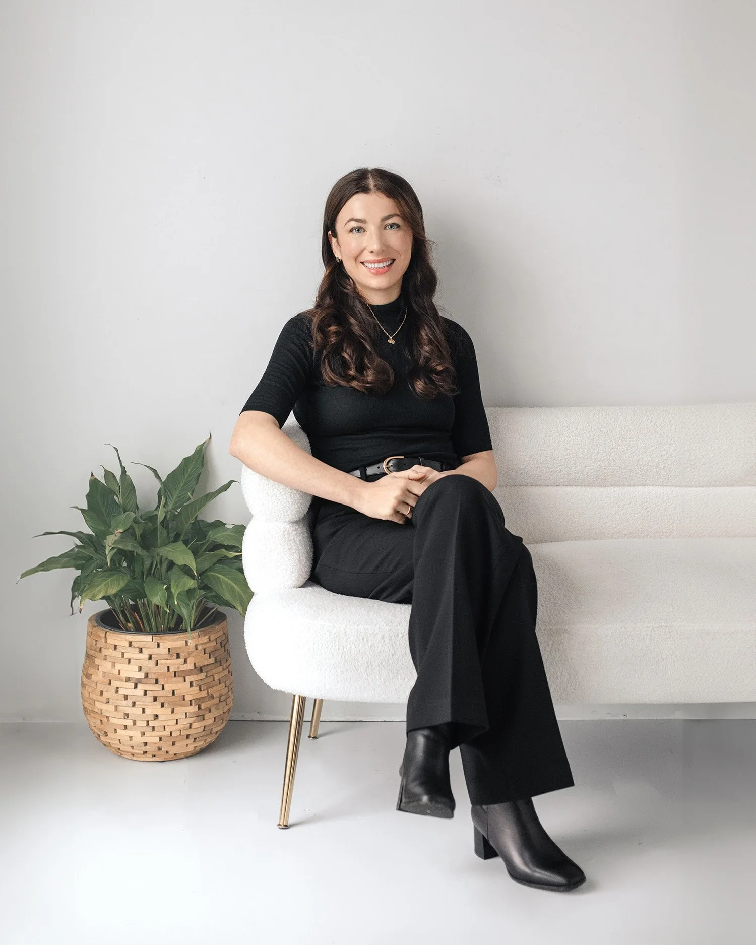 Portrait of psychologist Dr. Ivana Macar smiling, wearing a black outfit and seated in a chair beside a plant against a neutral wall.