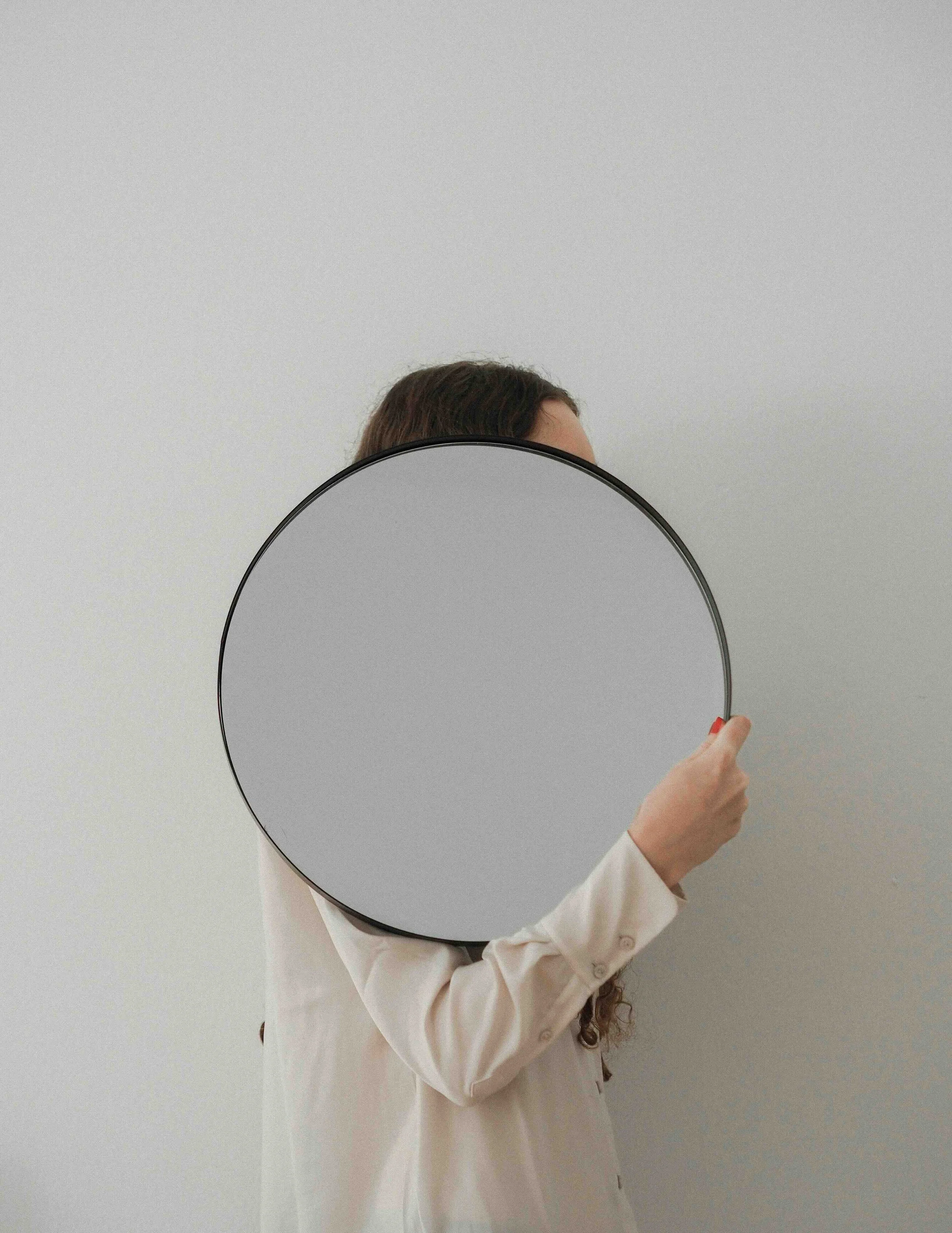 Person holding a round mirror in front of their face against a neutral wall.