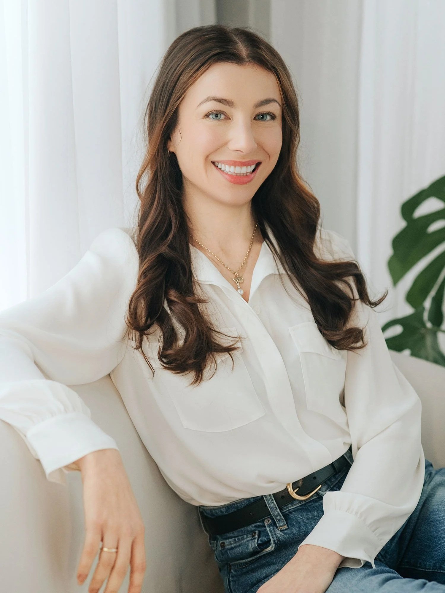 Portrait of Dr. Ivana Macar, psychologist, with long brown hair wearing a light blouse and necklace, seated and facing slightly toward the camera in a softly lit indoor setting with a neutral background and plant detail.