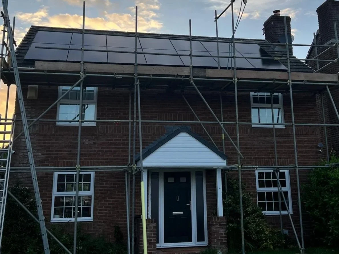 A two-story brick house with scaffolding around the front, and solar panels installed on the roof, with a cloudy sky in the background.