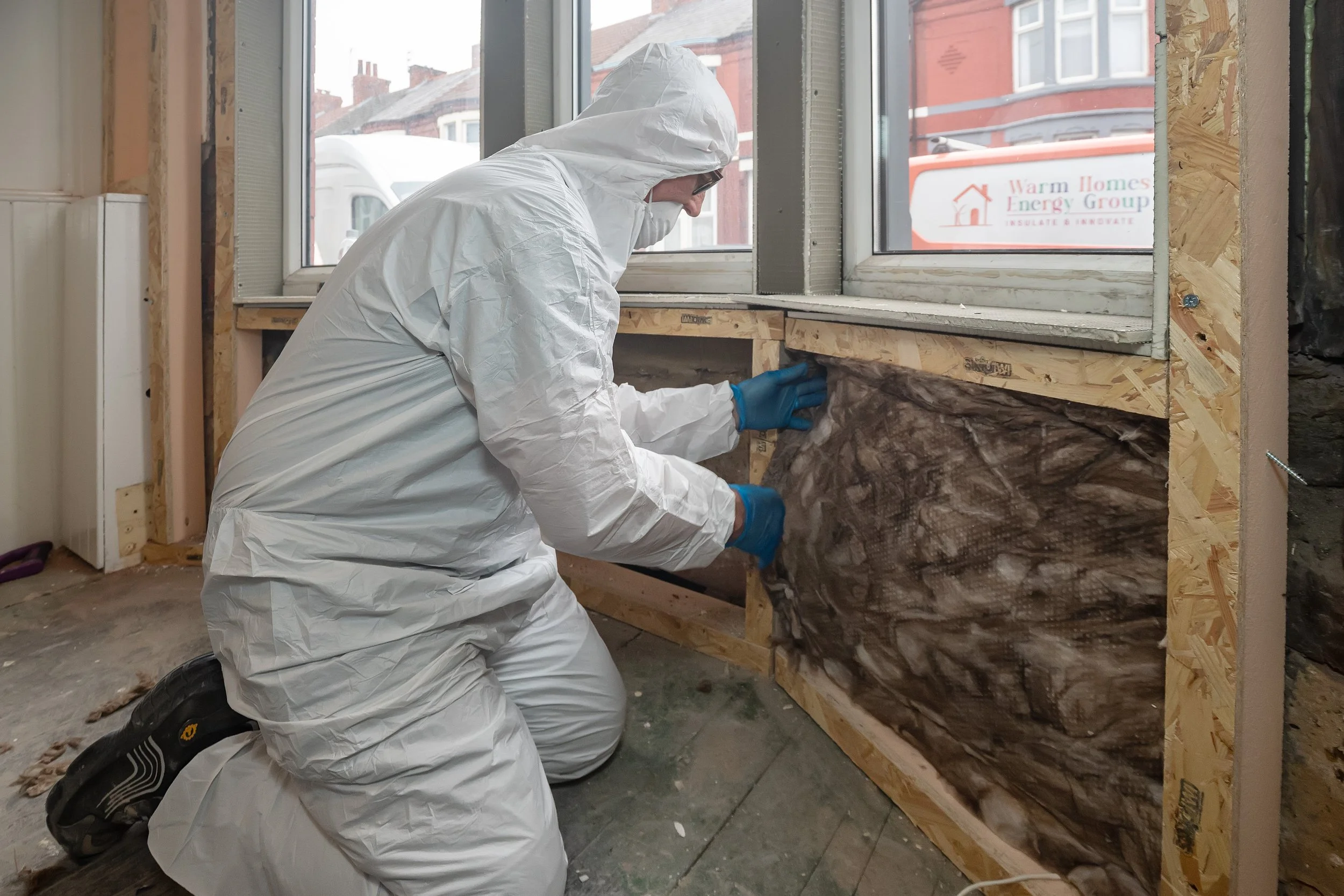 Person in protective suit installing insulation in a wall near a window in a building under construction.