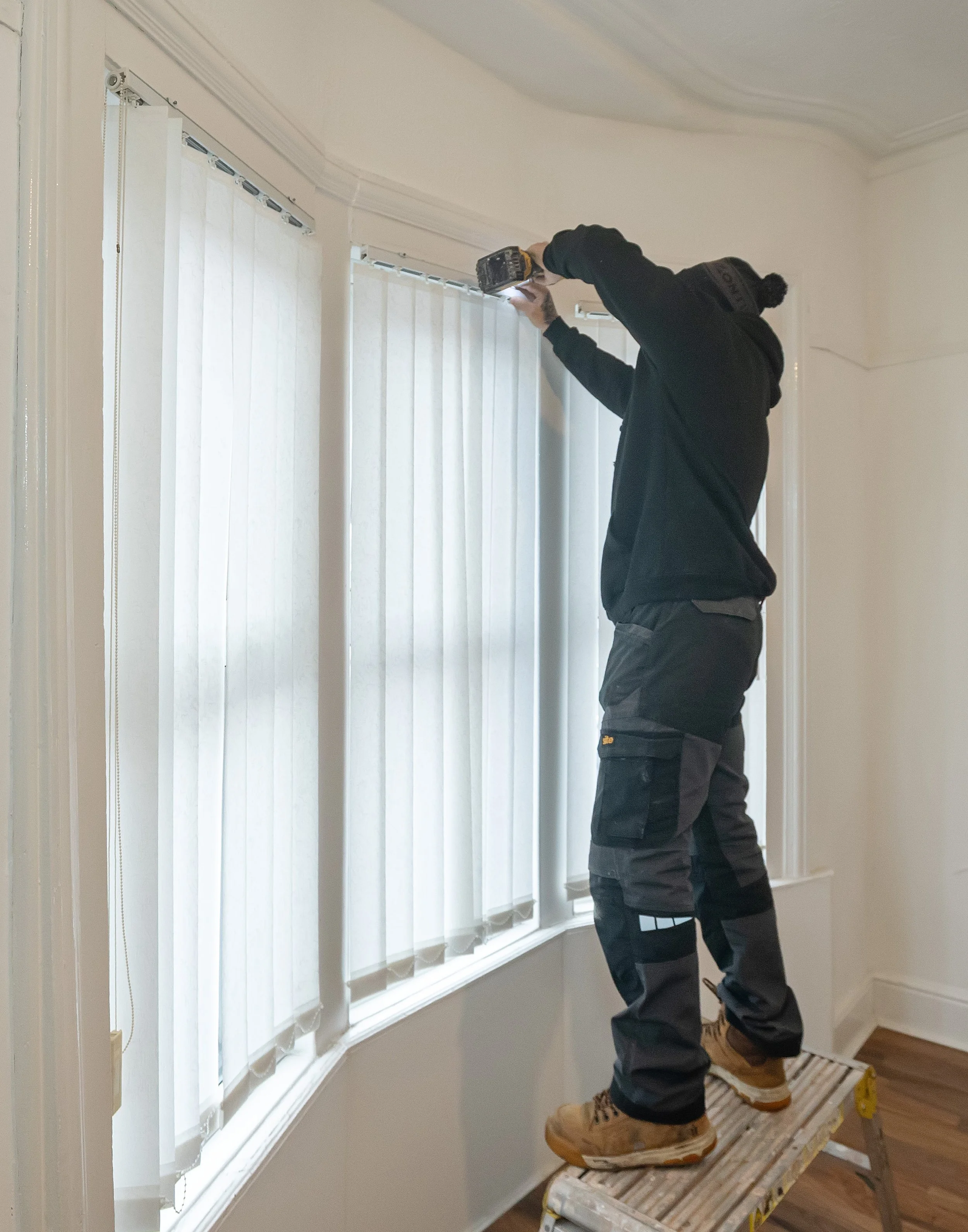 A worker wearing a black hoodie, dark work pants, and tan boots, standing on a ladder, installing or repairing window blinds with a cordless drill.