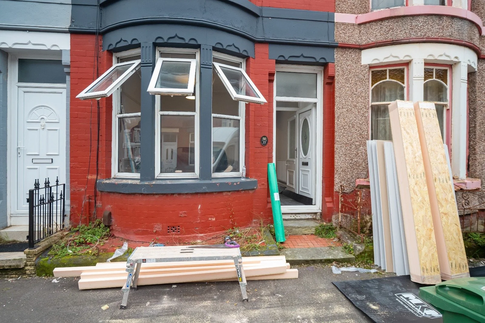 Front of a red brick house with a bay window and a white front door, with construction materials and tools outside indicating renovation work.