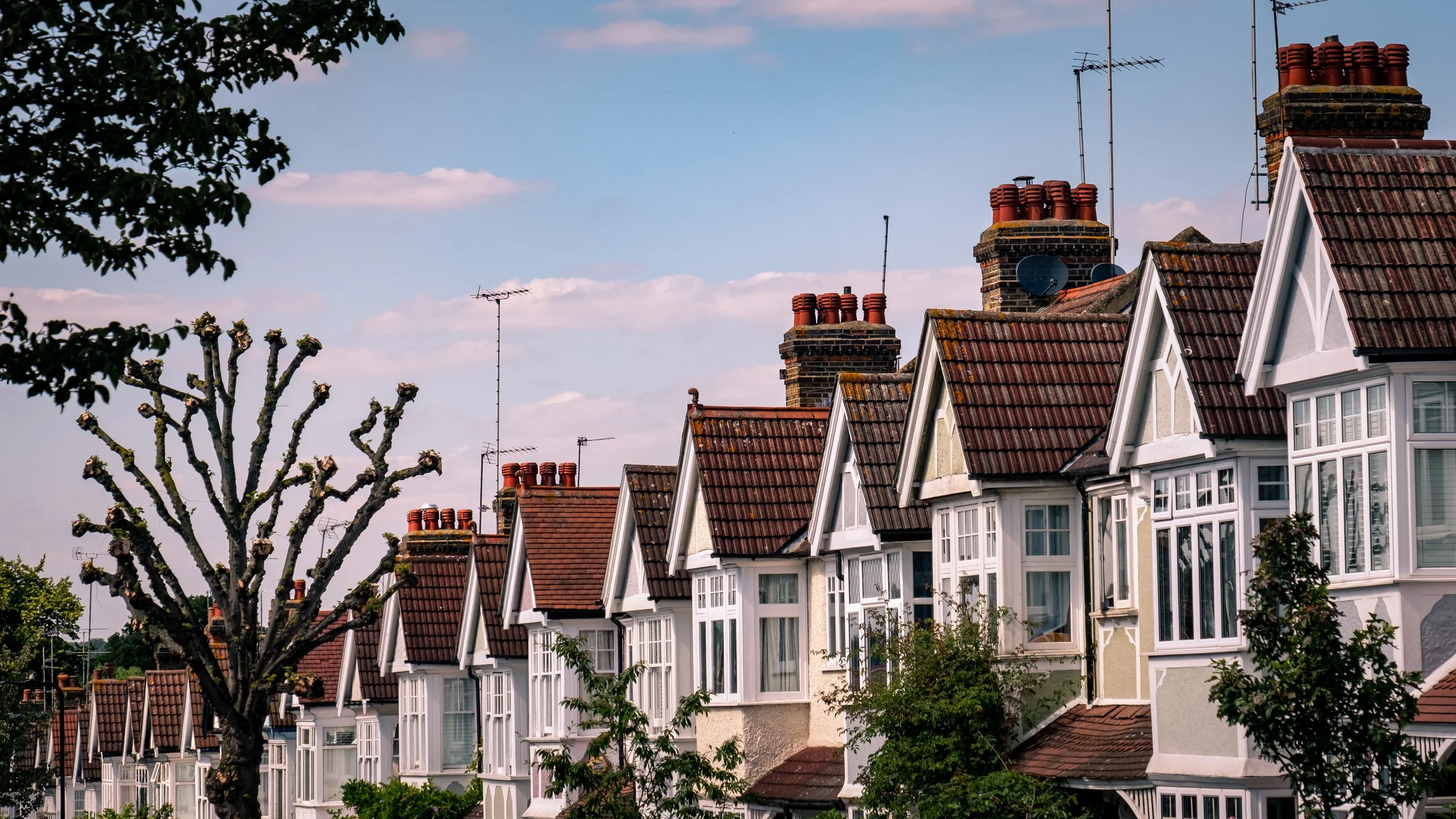 A row of suburban houses with white and yellow exteriors, steep red tile roofs, and multiple chimney stacks against a blue sky with scattered clouds. Some trees are visible in the foreground.