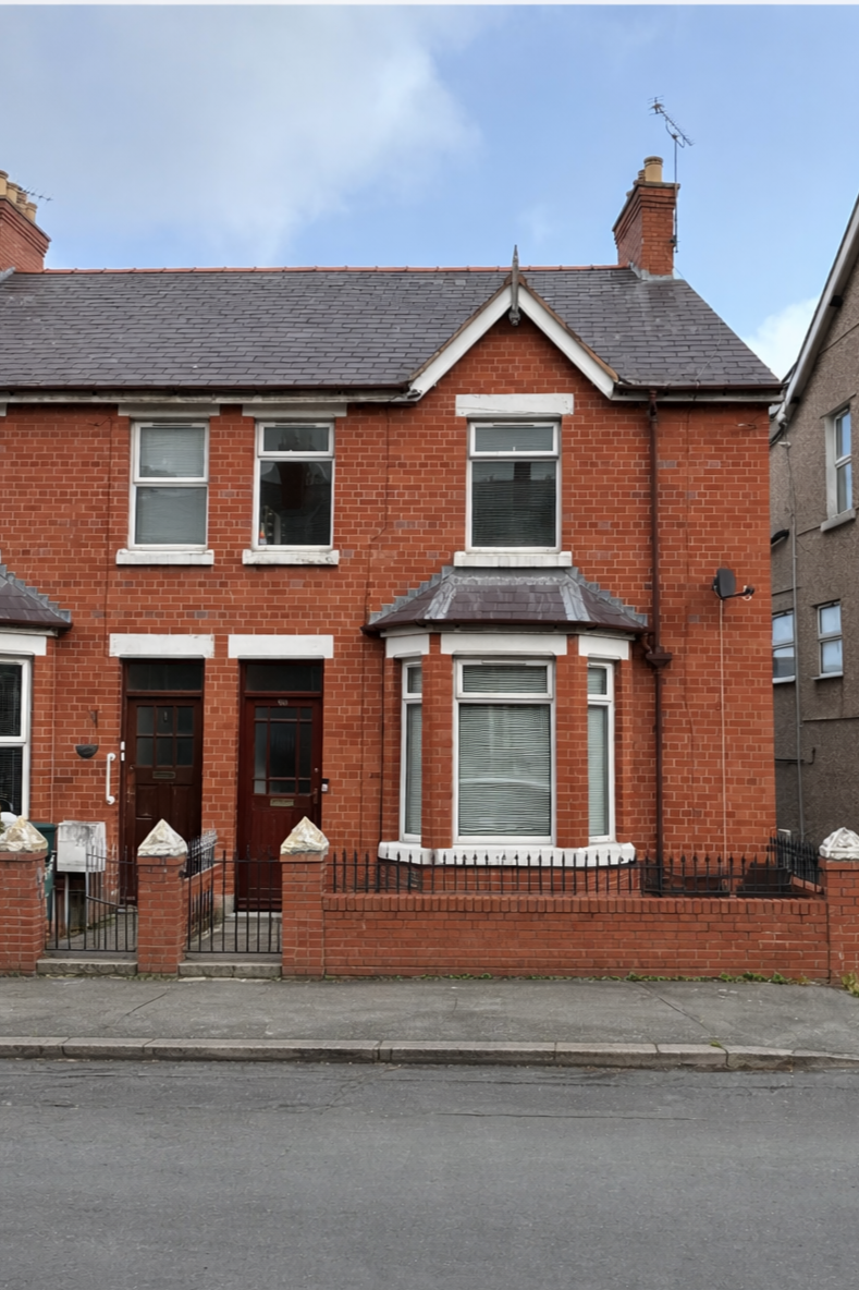 A brick house with three stories, featuring a small front yard with a brick fence and a sidewalk. The house has multiple windows with white trim and a small porch area. The roof is gray, and there is a chimney with an antenna on top.
