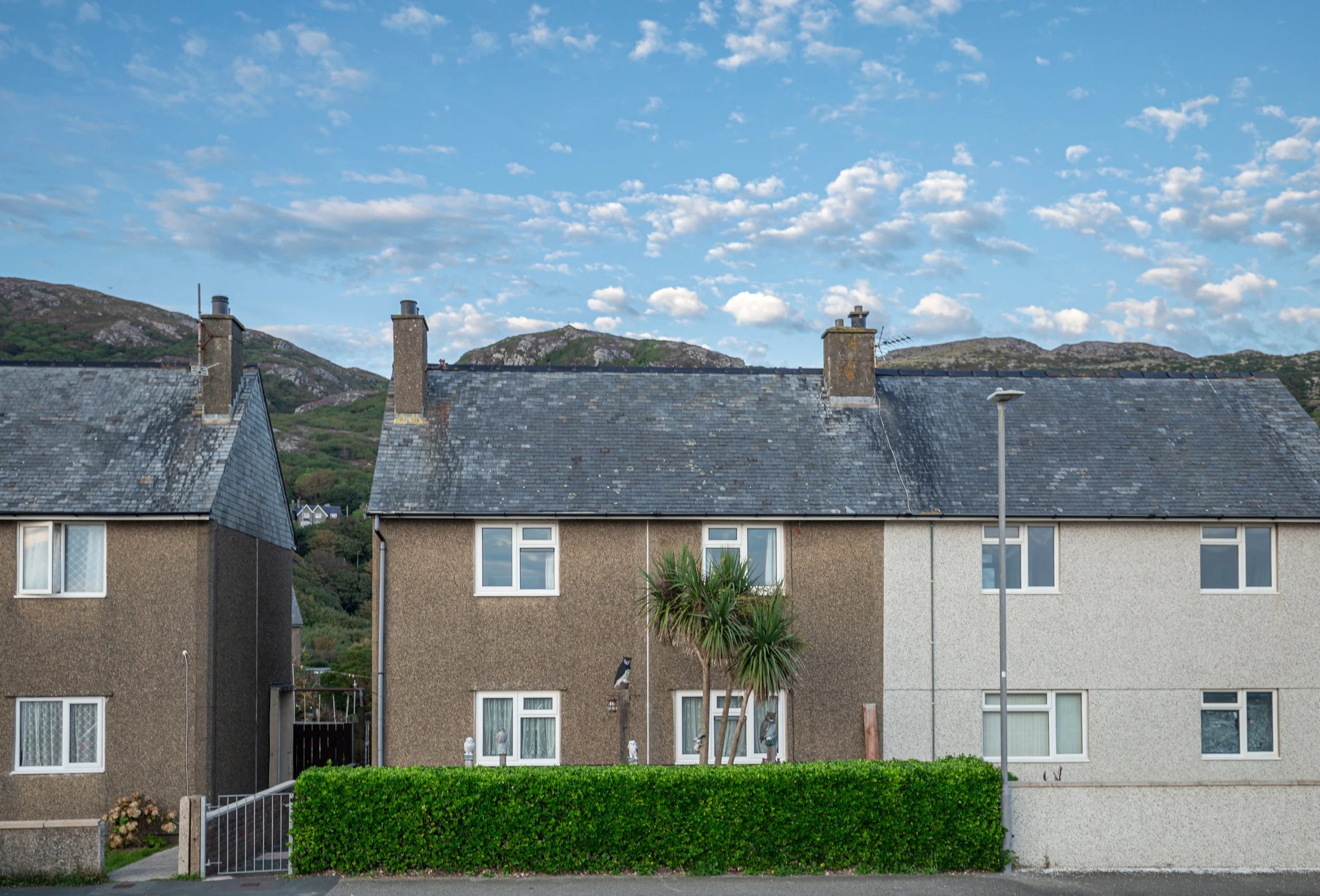 Three houses with gray roofs and small windows, a hedge in front, and a palm tree, under a partly cloudy sky with mountains in the background.