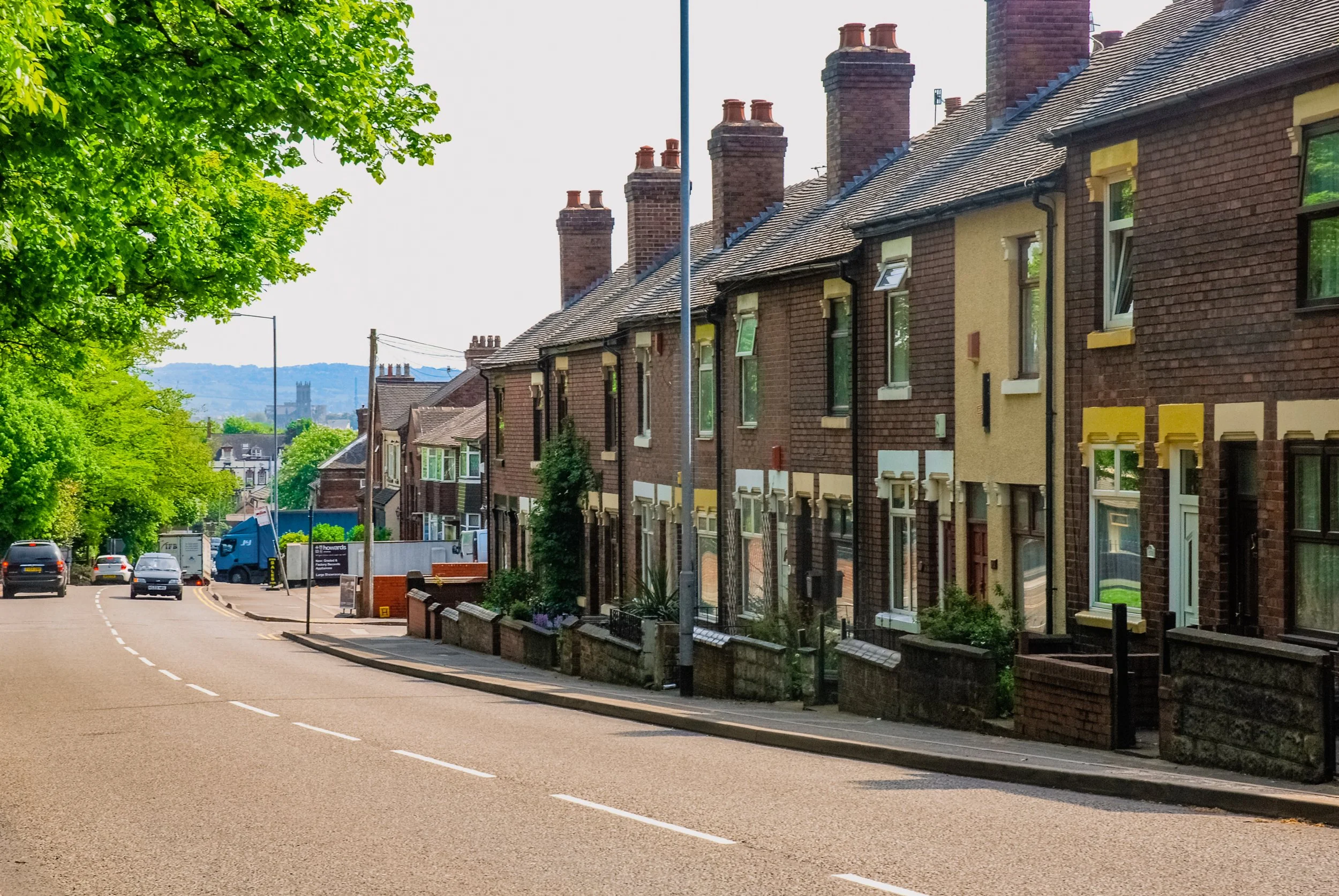 A residential street with brick houses, trees on the left, and cars parked along the road. The houses have chimneys and small front yards.