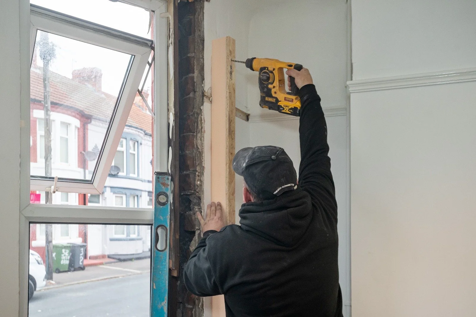 A person is using a cordless drill to work on a wall near a window, with a level tool propped against the wall.