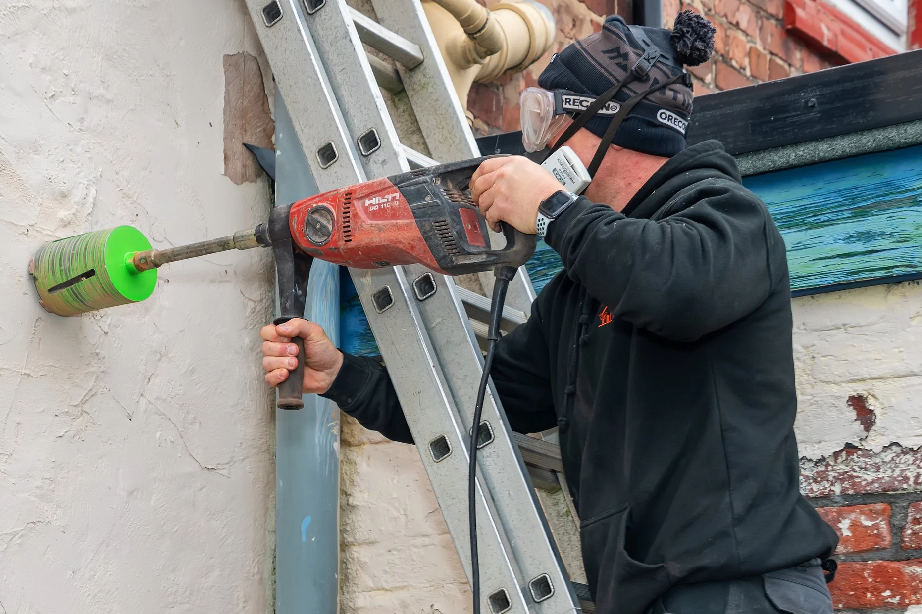 A worker wearing a black hoodie, safety goggles, and a beanie is using a power drill to work on a wall. The wall has a brass pipe with a green can over it and a ladder is positioned nearby.