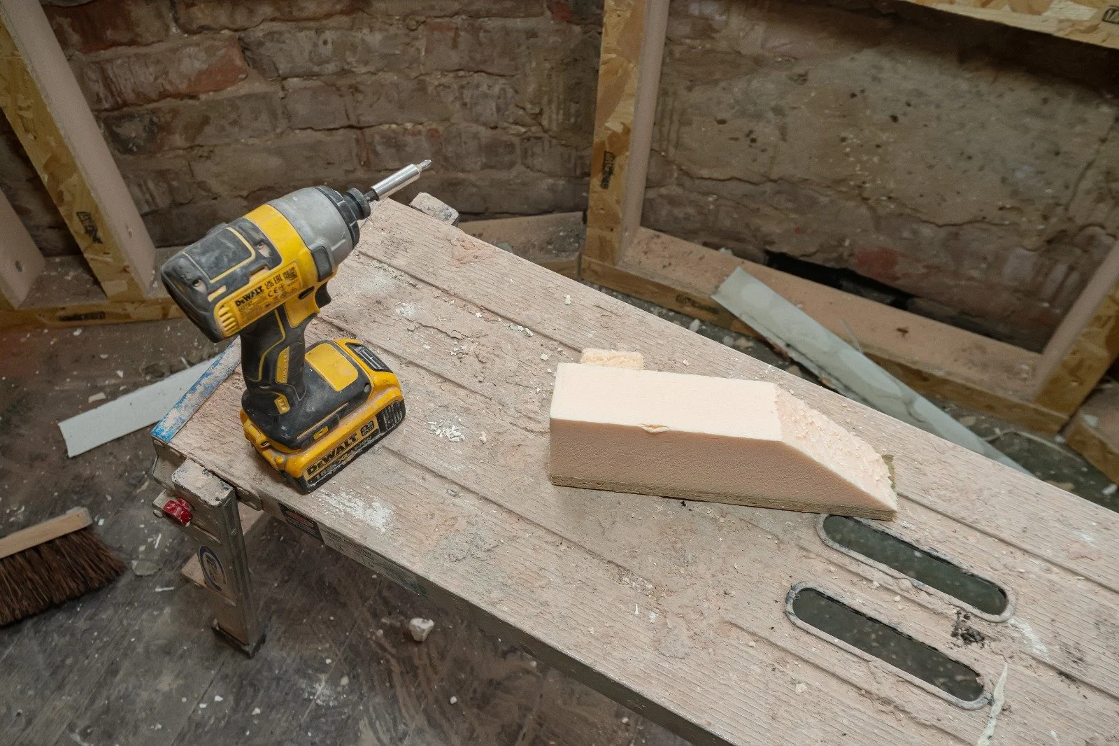 A woodworking workbench with a yellow cordless drill and a wooden block on it, in a construction site setting with brick and wood framing in the background.