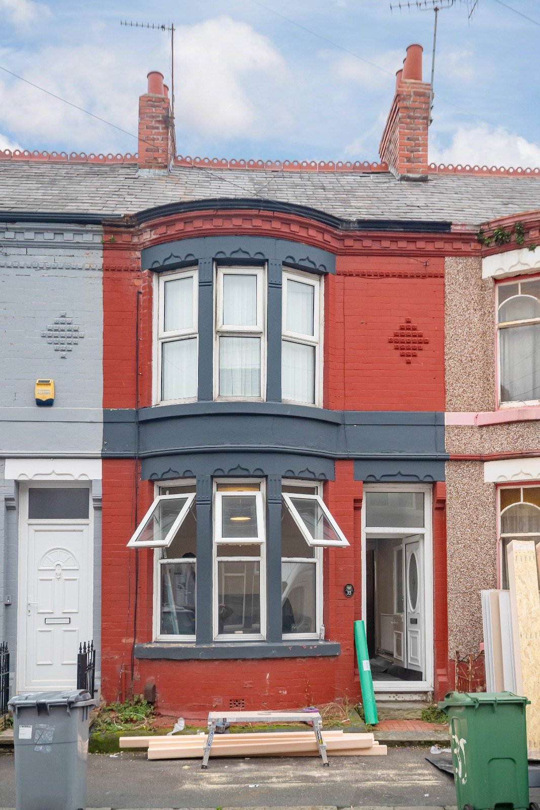 A colorful row house with a blue and red facade, featuring bay windows and a front door, under renovation with building materials and tools outside.