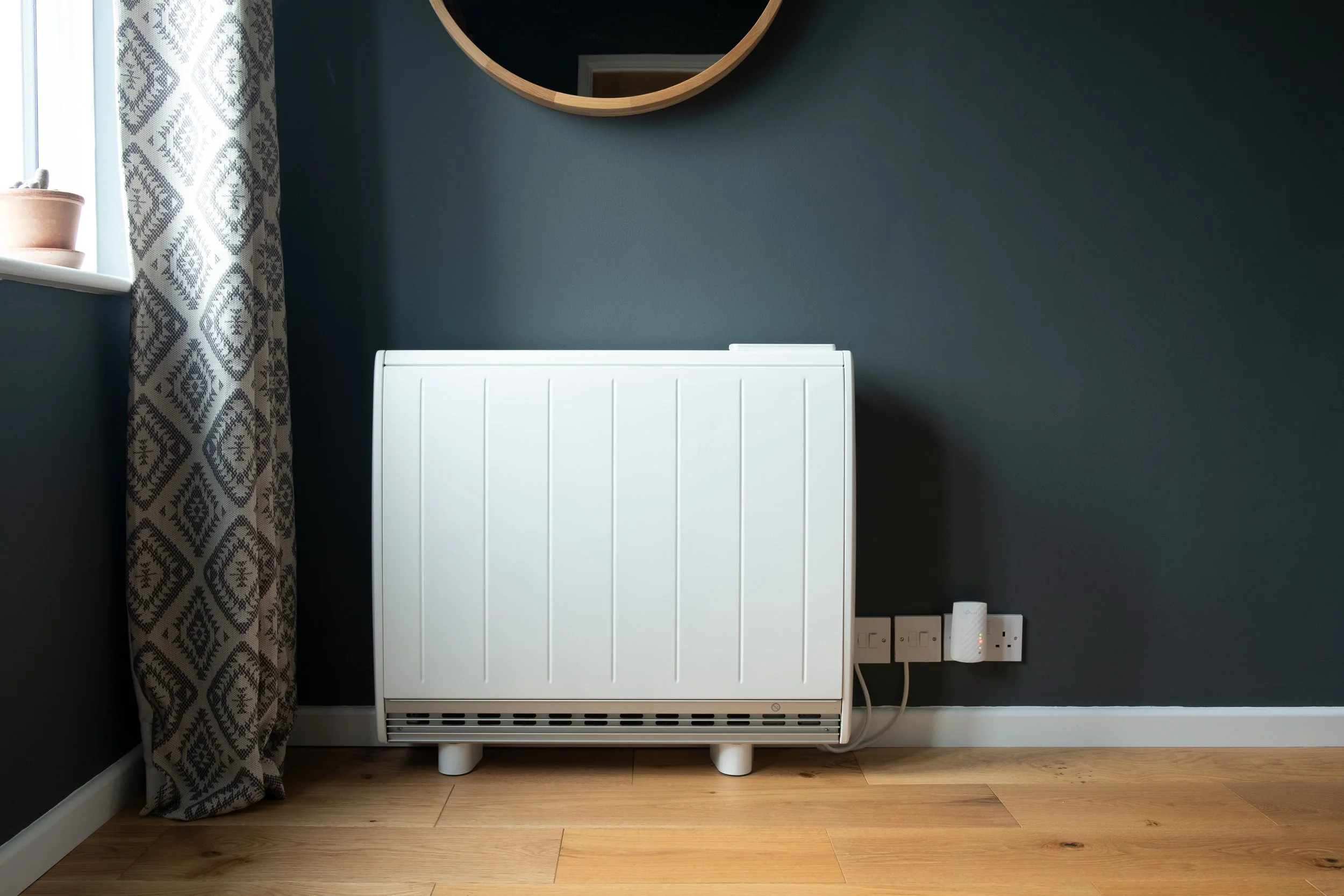 A white radiator against a dark green wall with a round wooden mirror above it, a window with patterned curtains on the left, and electrical outlets on the right.