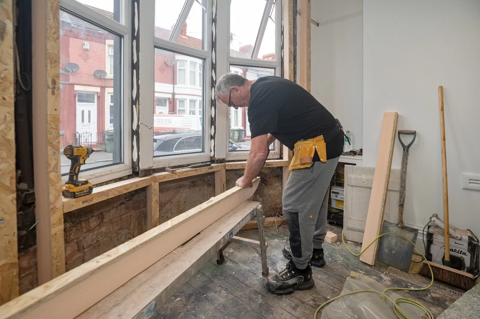 A man working on a construction project inside a room with large windows, using a saw to cut a piece of wood placed on a workbench. Various tools and construction materials are visible around him.