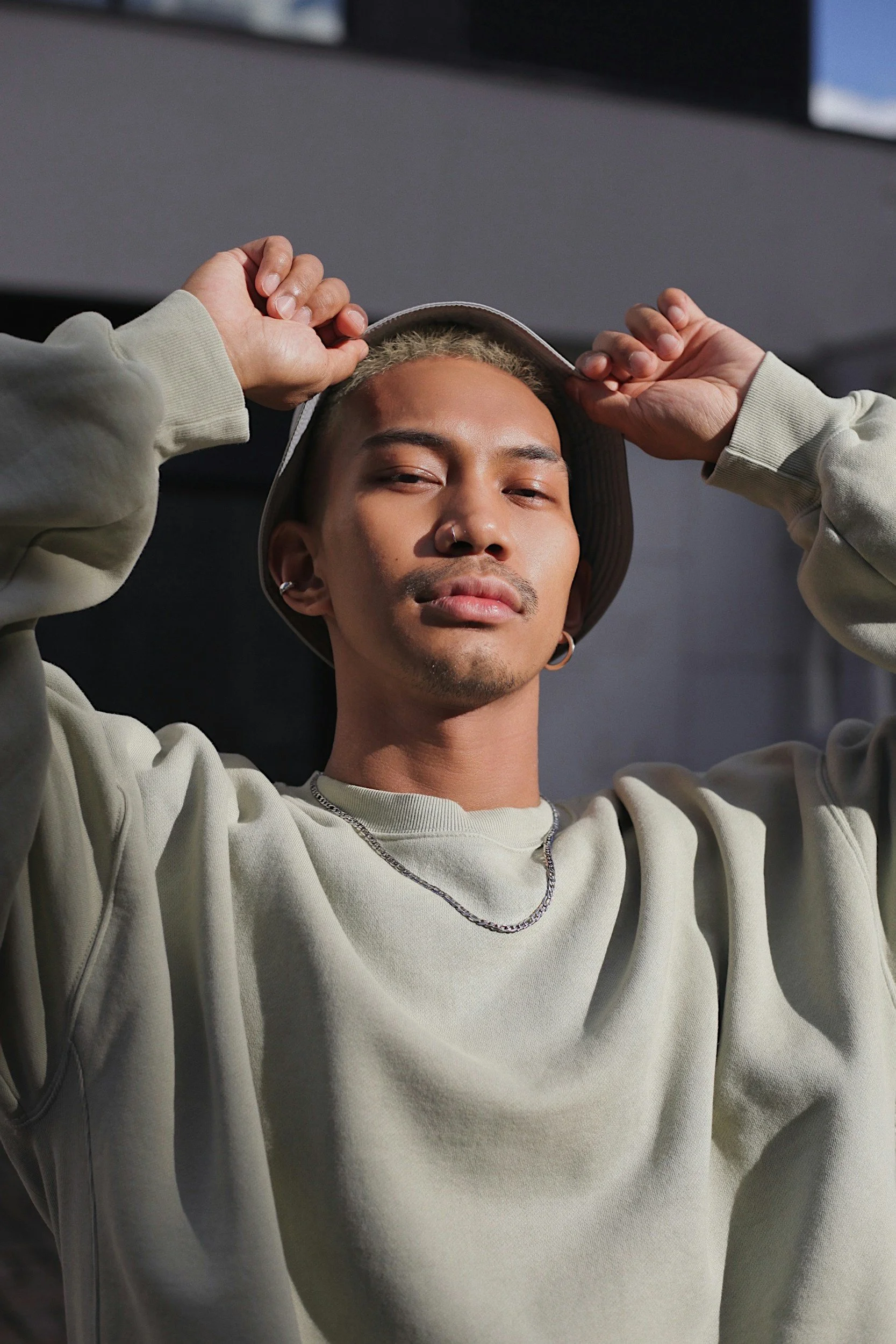A person with a contemplative expression poses with hands behind their head, wearing a light sweater and a hat. The background is softly lit, giving a relaxed vibe.