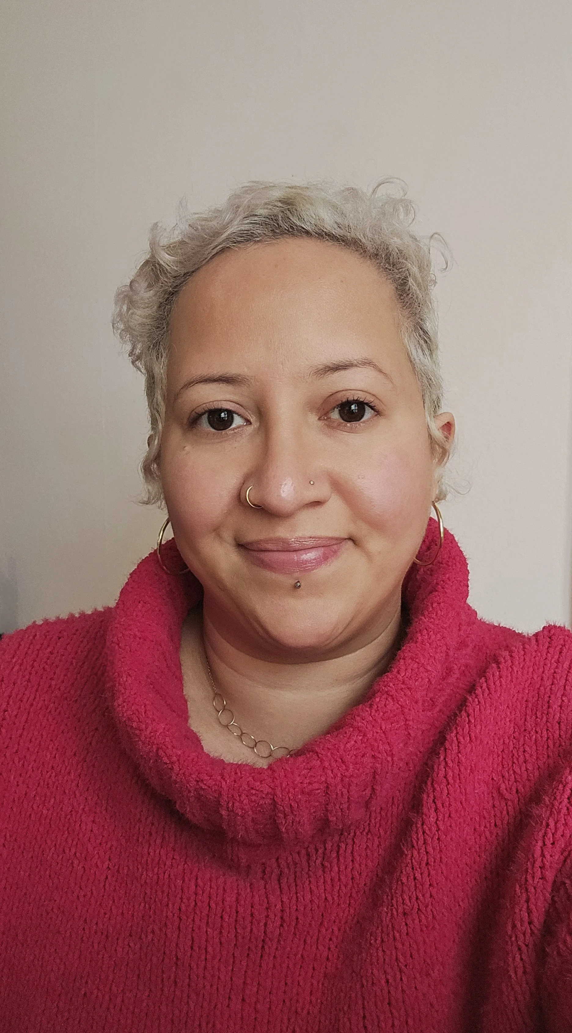 A person with short, curly blonde hair and hoop earrings smiles warmly. They wear a bright pink sweater, exuding a friendly and inviting demeanor.