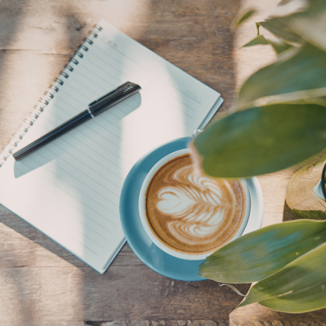 A notebook and pen beside a blue saucer holding a latte with leaf latte art. Sunlight and green leaves create a calm, inviting atmosphere for a therapy session.