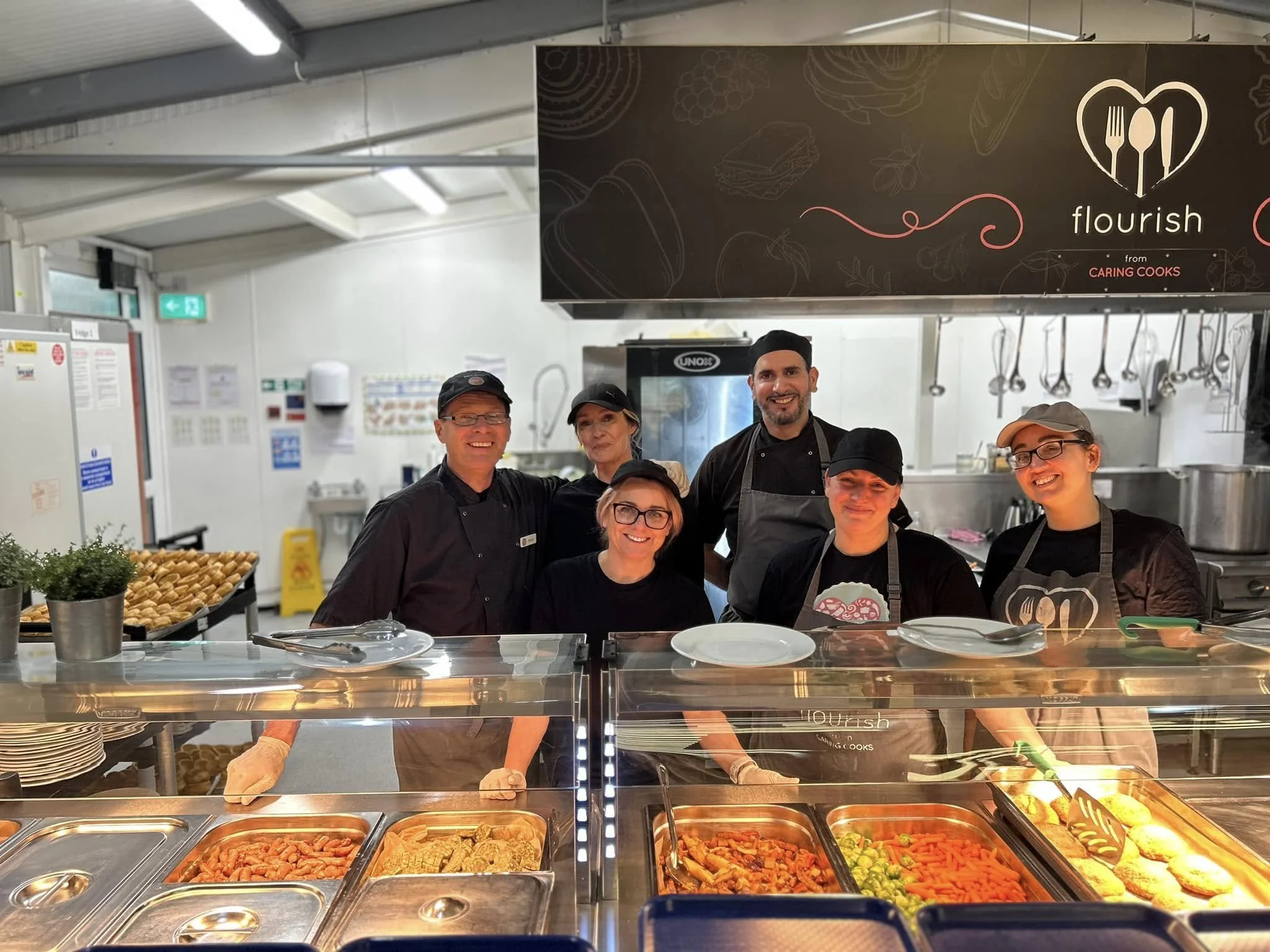 Group of six smiling kitchen staff members standing behind a food counter in a professional kitchen, with a sign reading 'flourish from caring cooks' above them.