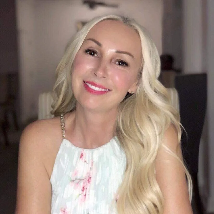 A woman with long blonde hair smiling while sitting indoors.