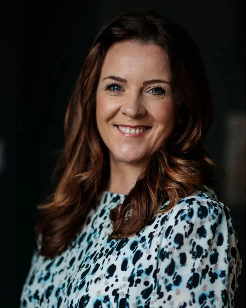 A woman with long, wavy, auburn hair, blue eyes, and light skin, smiling at the camera. She is wearing a white blouse with black and blue leopard print patterns. The background is dark and blurred.