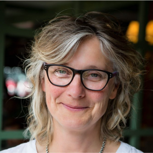 Close-up portrait of a woman with wavy blonde hair, wearing glasses, smiling outdoors with blurred background.