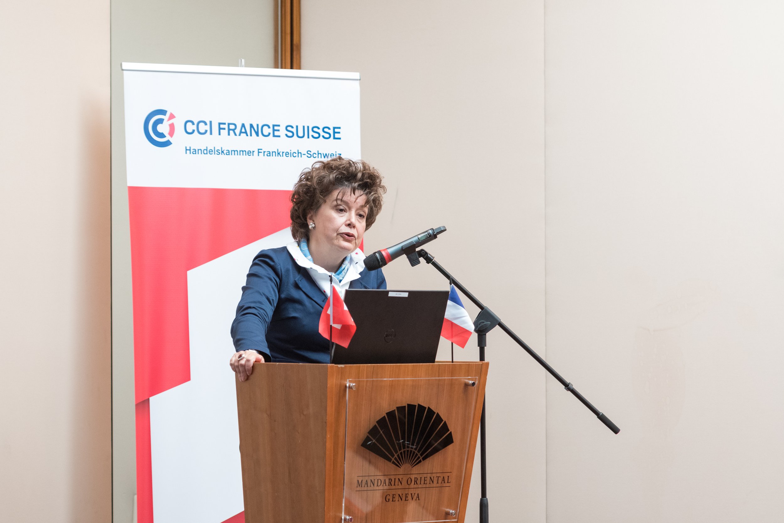 Femme à une conférence, debout derrière un pupitre en bois portant deux petits drapeaux, parlant dans un micro, avec un panneau affichant 'CCI FRANCE SUISSE' en arrière-plan.