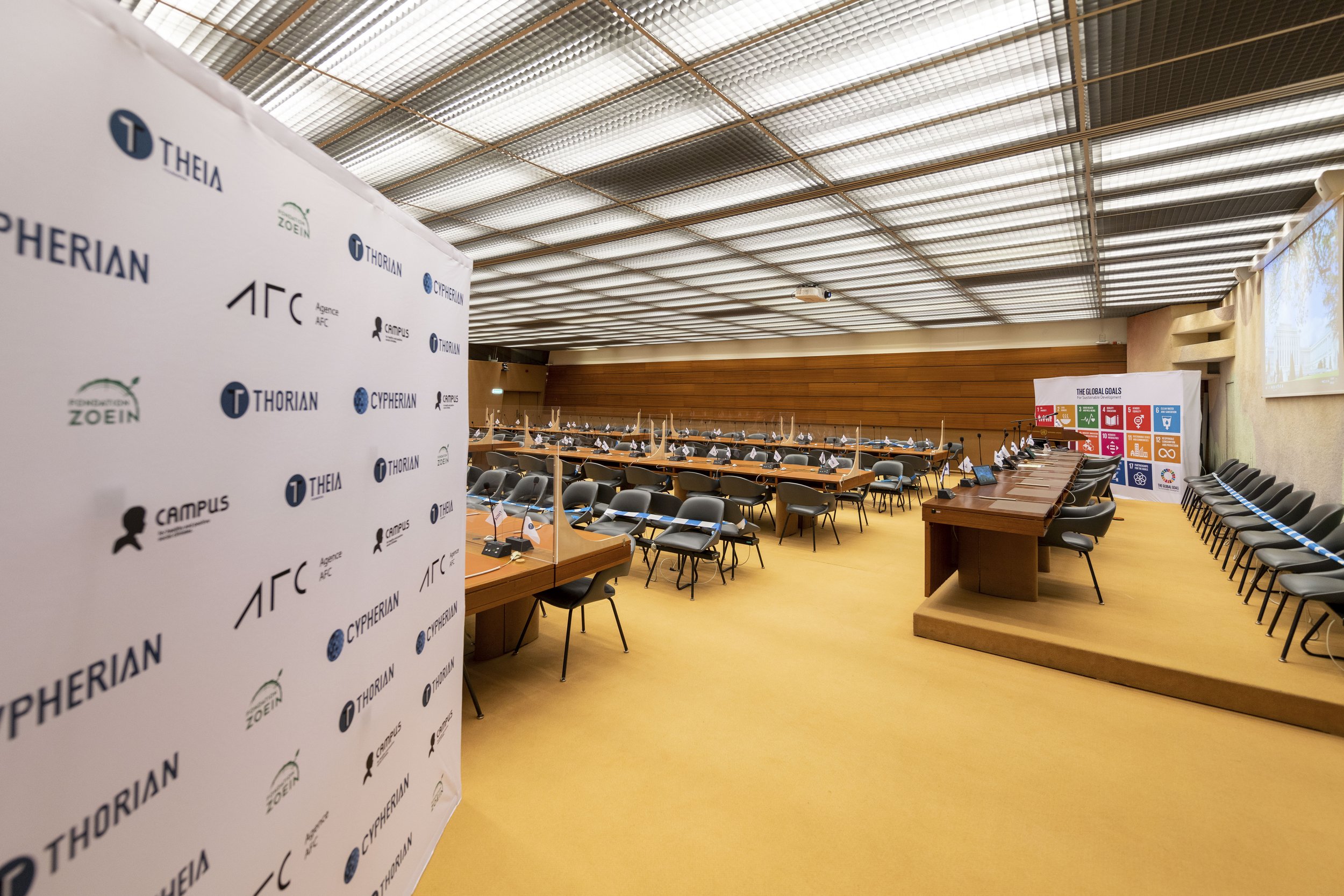 Salle de conférence avec des tables, des chaises et des panneaux de séparation en plastique, décorée avec des affiches sur les objectifs de développement durable et des logos de différentes organisations.