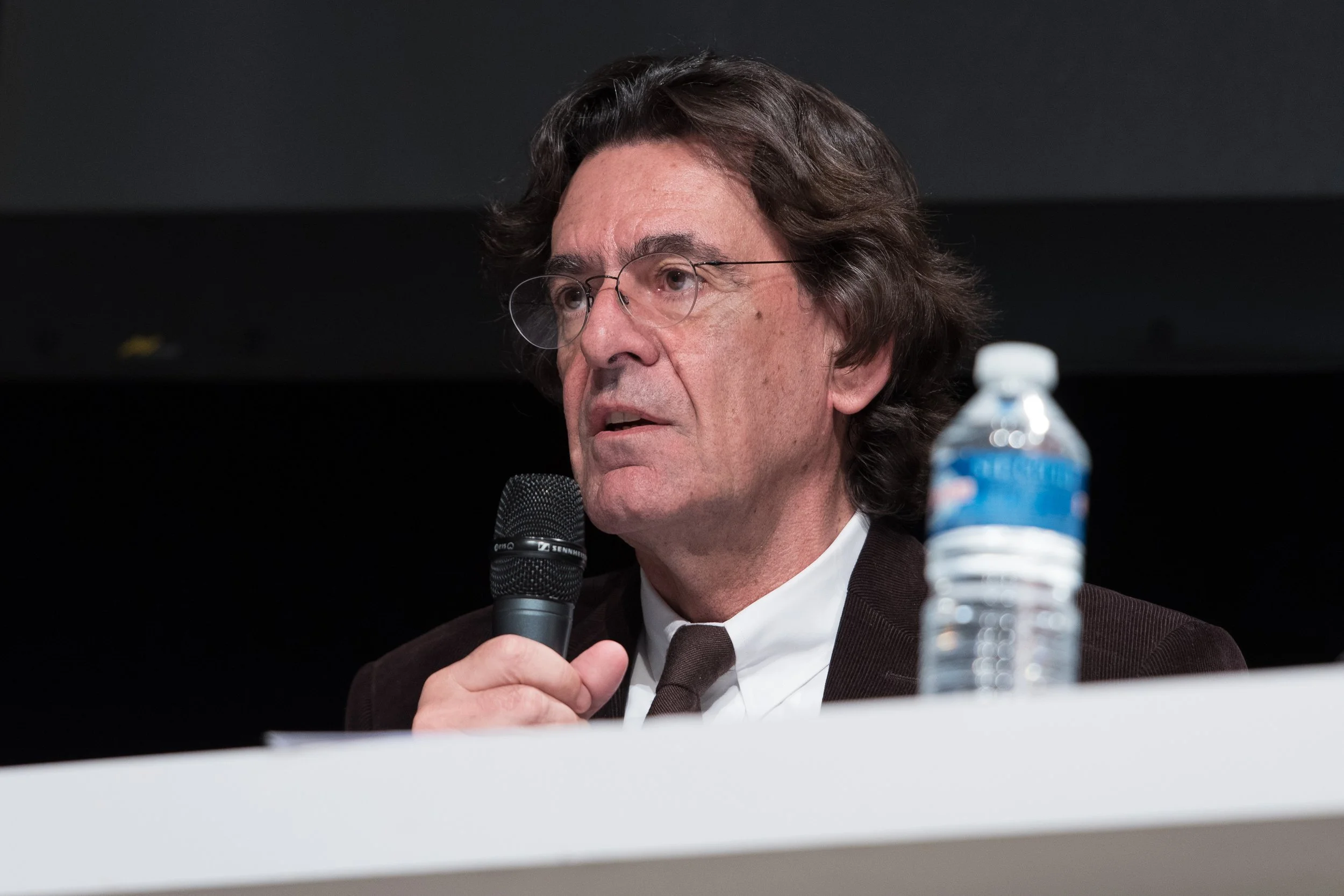 Un homme portant des lunettes, un costume sombre, et une cravate, tient un micro lors d'une conférence ou d'une panel discussion.