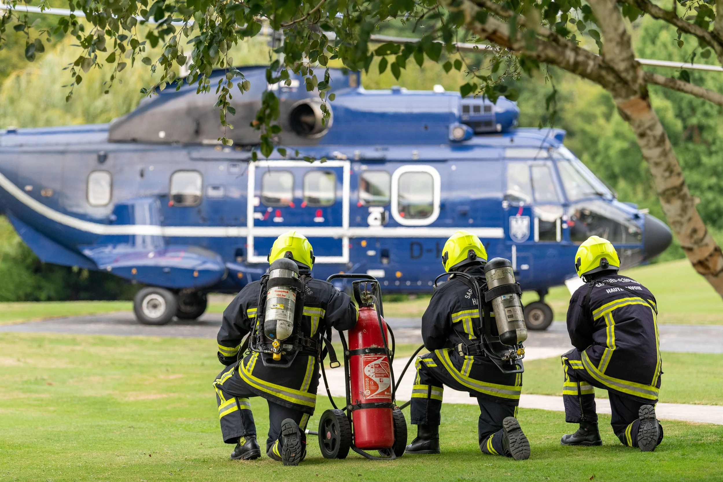 Trois pompiers en tenues de protection en position agenouillée sur l'herbe, équipés de bouteilles d'oxygène, regardant un hélicoptère de couleur bleue posé sur le sol, avec des arbres en arrière-plan.