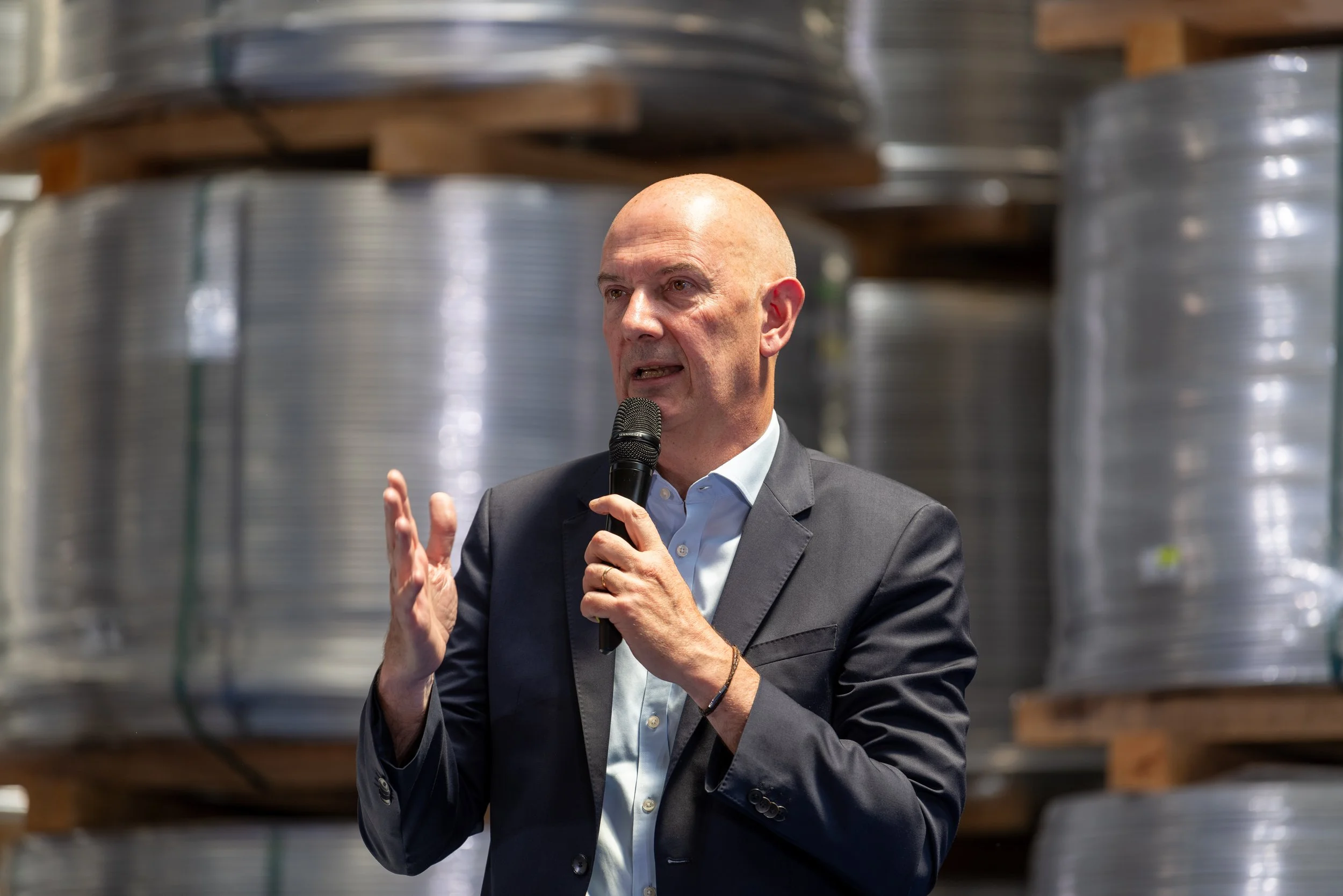 Homme parlant dans un micro lors d'un événement, en arrière-plan des bobines de fil industriel.