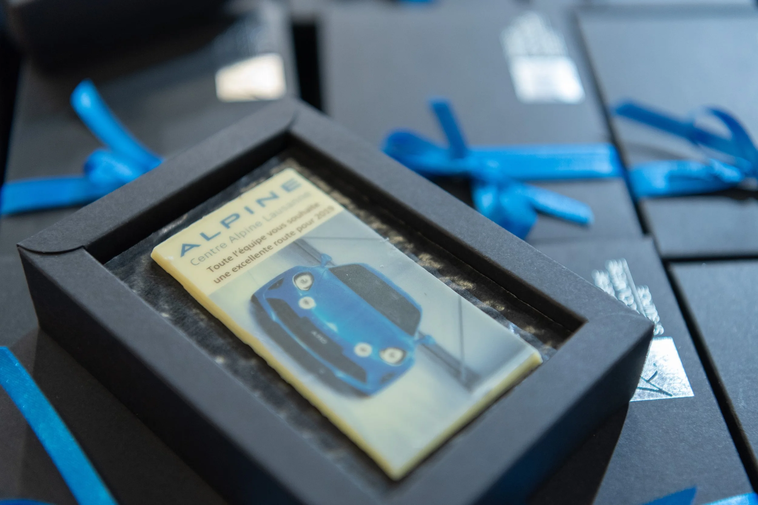 Une plaque cérémonielle avec une image d'une voiture bleue et le texte 'ALPINE' et un message de félicitations, entourée de boîtes noires avec des rubans bleus.