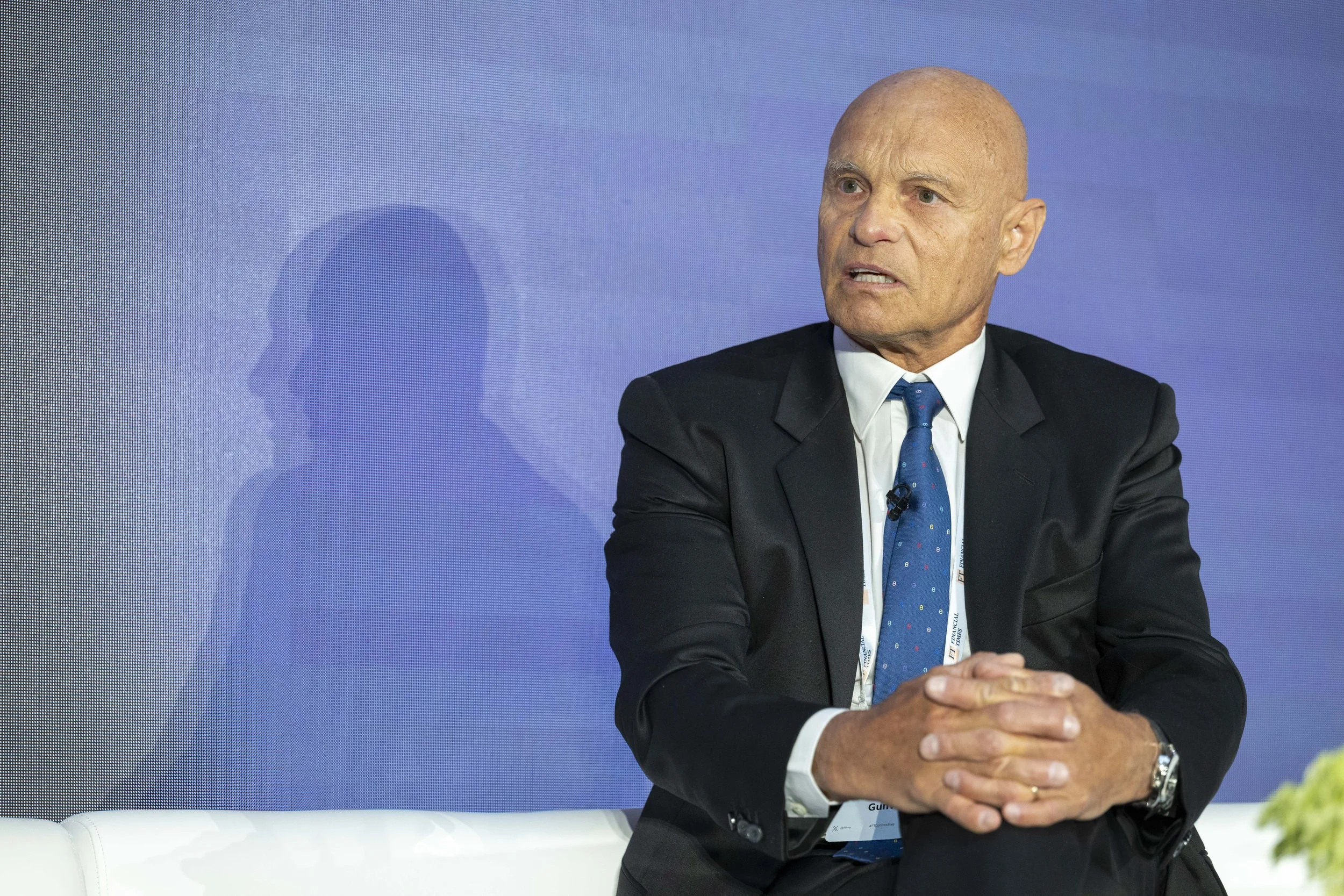 Un homme d'affaires en costume noir, blanc chemise et une cravate bleue, assis sur une scène lors d'une conférence ou d'un panel.