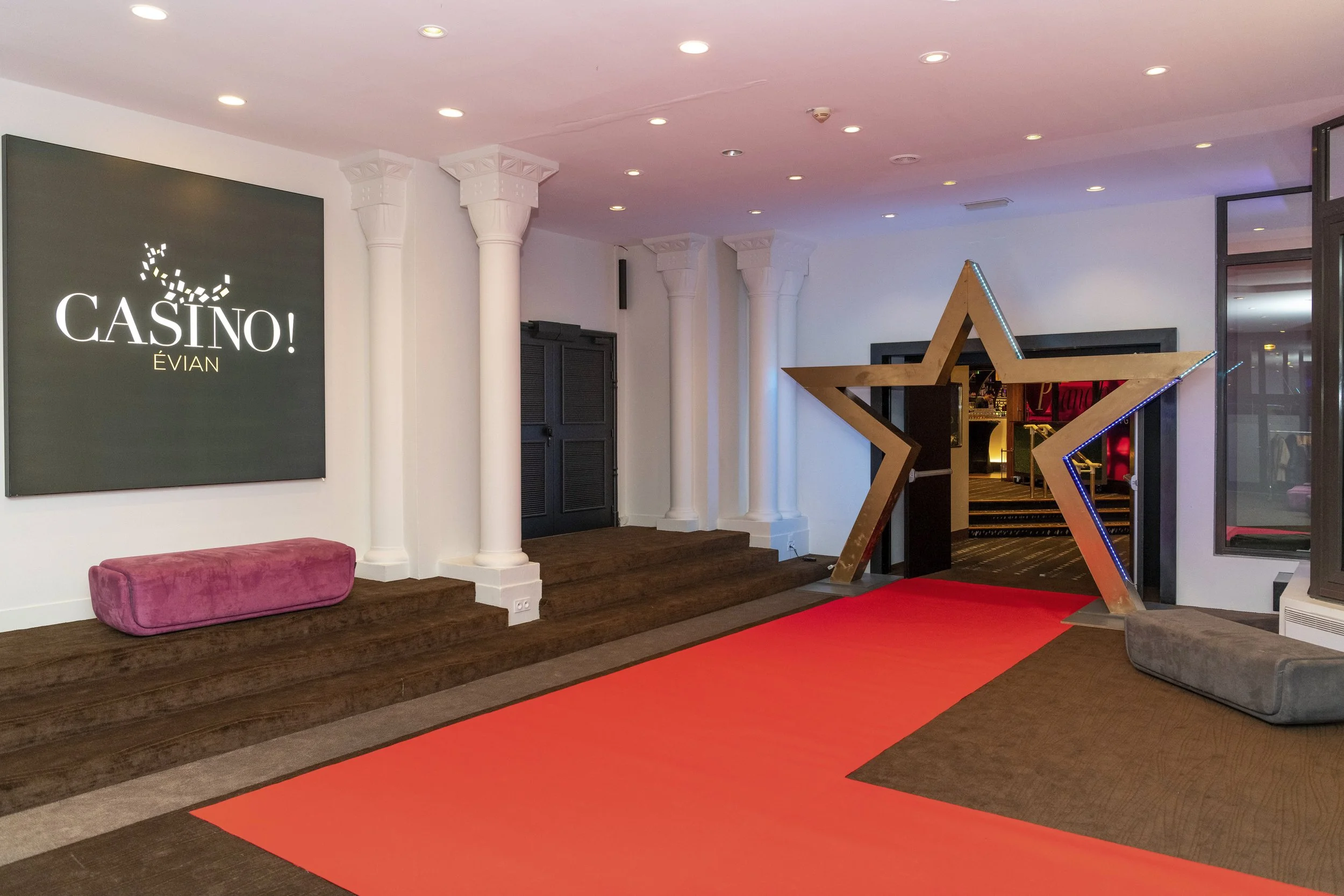 L'entrée d'une salle de casino avec un tapis rouge, une étoile en bois stylisée à l'entrée, et un panneau indiquant 'CASINO! ÉVÍAN' sur un mur blanc.